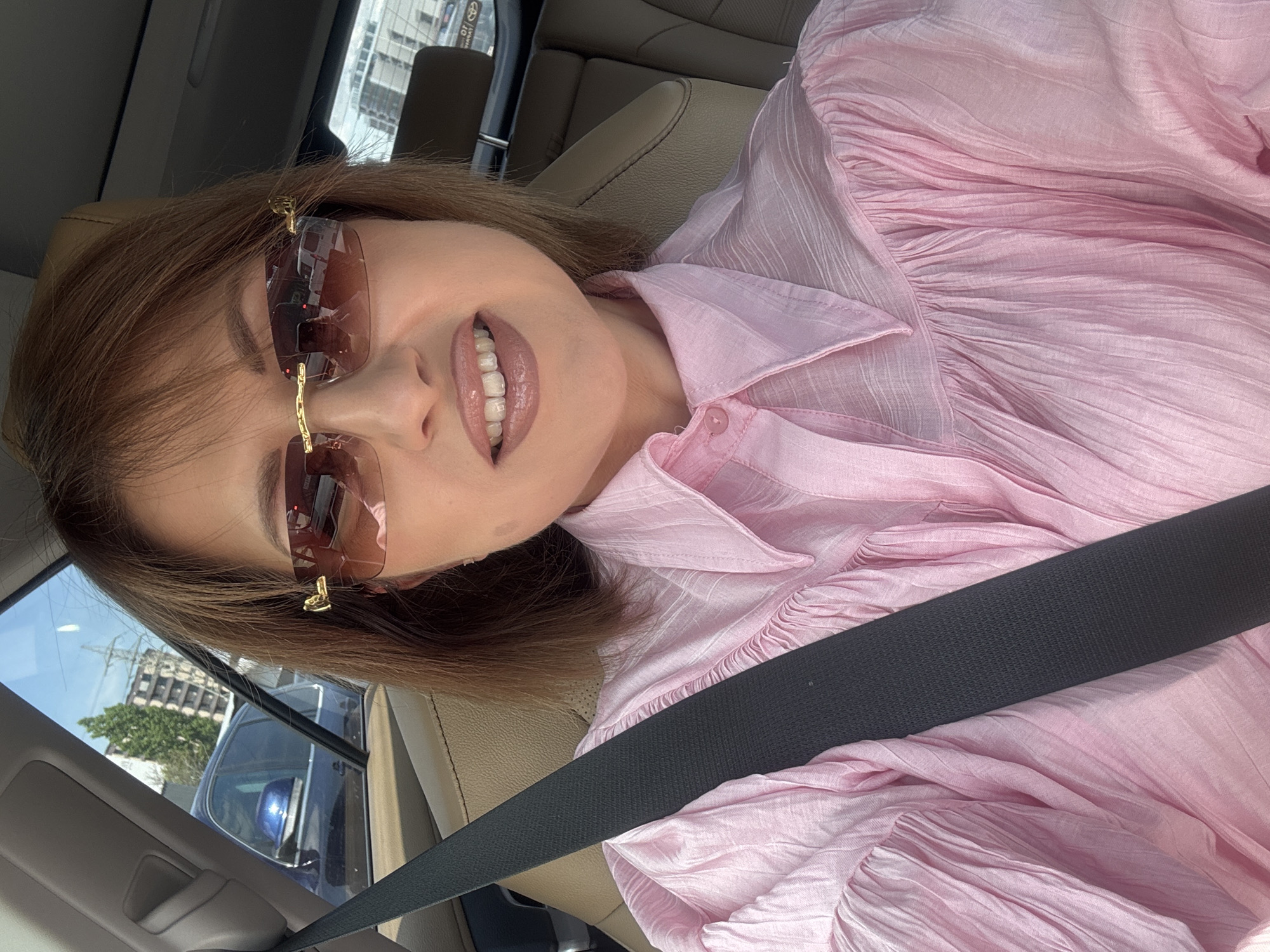 A medium shot depicts a woman wearing a pink shirt and sunglasses, seated in a car. She is wearing a seatbelt. The car's interior is beige. Through the car window, a cityscape can be seen, with buildings and vegetation visible. The image was taken in Sofia, Bulgaria, and shows the interior of a Toyota automobile.