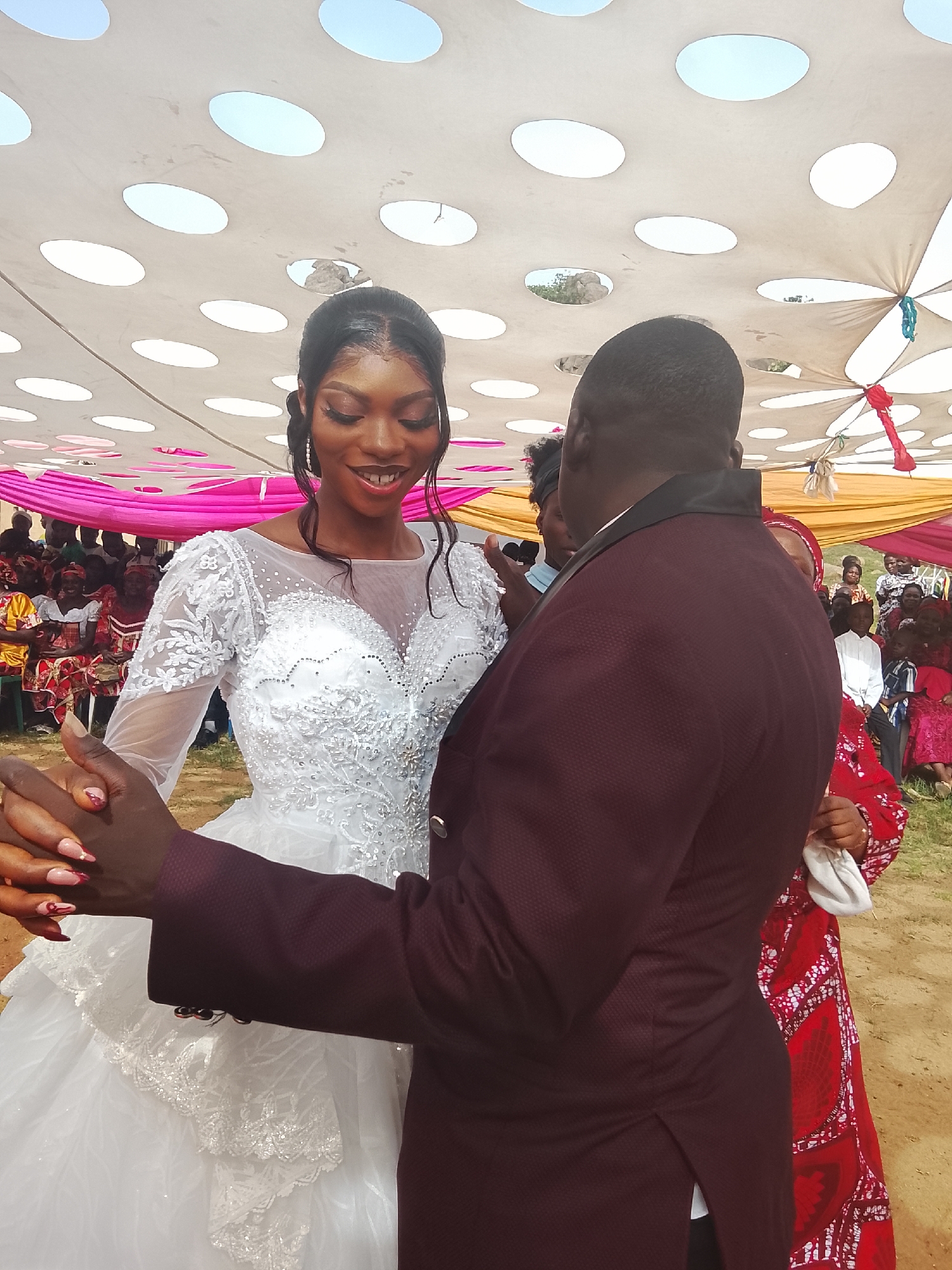 A photograph taken outdoors in Dogon Dinia, Nigeria, shows a bride and groom during a wedding celebration. The bride, wearing a white, full-length gown with a sheer neckline, lace embellishments, and a layered skirt, is seen from the waist up. She has dark hair styled back from her face and is smiling as she looks towards the right side of the frame. The groom, whose back is predominantly to the camera, is wearing a dark burgundy suit jacket. The groom's hands are holding the bride's hands. Numerous wedding guests can be seen in the background; most are seated. Above the group is a canopy tent. The ground appears to be dirt or sand.