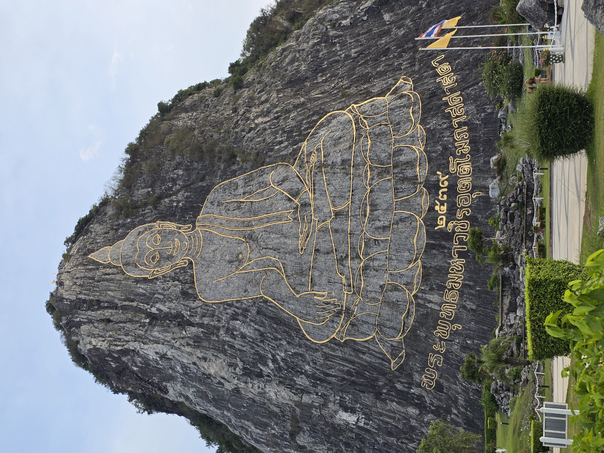 The image shows a large Buddha image carved into a rocky mountainside in Tambon Bang Sare, Thailand. The Buddha is depicted seated in a cross-legged position, with gold-colored lines outlining its form against the gray rock. Below the Buddha image are inscriptions in Thai script, also in gold. Two flags, one yellow and one a combination of blue, white, and red, fly on poles to the right of the inscription. The lower part of the image shows a landscaped area with green bushes, trees, and paved pathways. Two people are visible walking in the distance near the flags. The sky above is a pale blue with some wispy clouds.