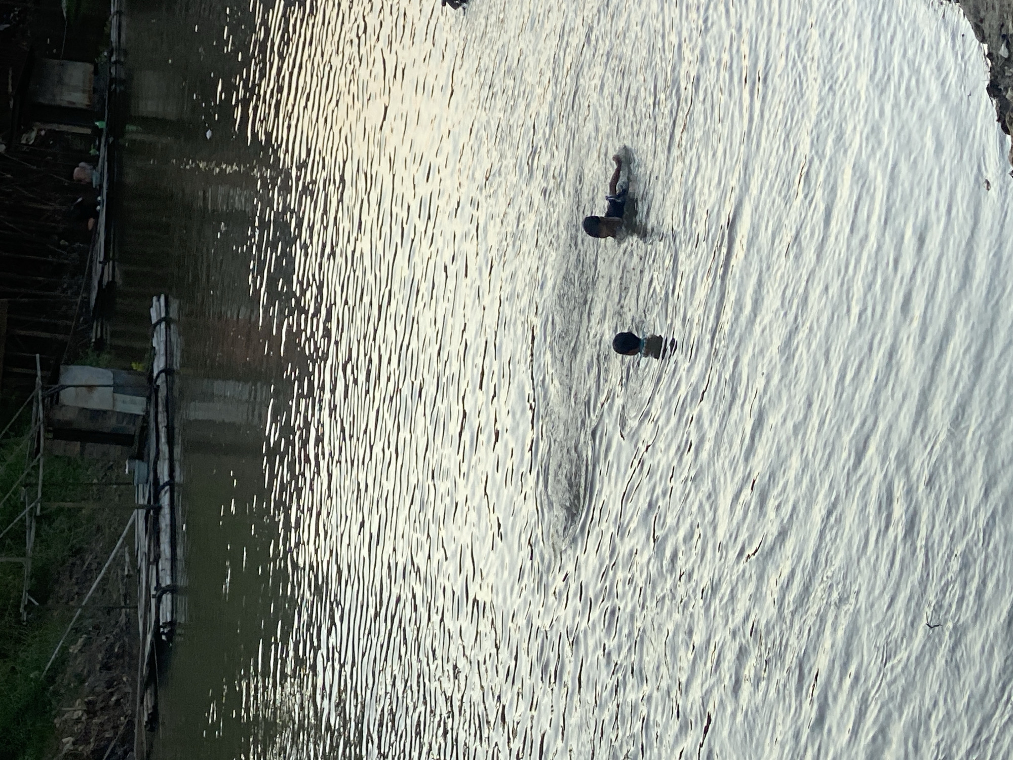 The image shows a body of water with two children swimming. In the background, there are structures built on stilts over the water, suggesting a riverside village or settlement. The water has ripples and reflections of light, indicating it is an open-air environment. The overall scene appears to be in a natural or semi-natural setting, possibly a river or a large pond, with evidence of human habitation along the banks. The location is identified as Tunggul Irang Ulu, Indonesia.