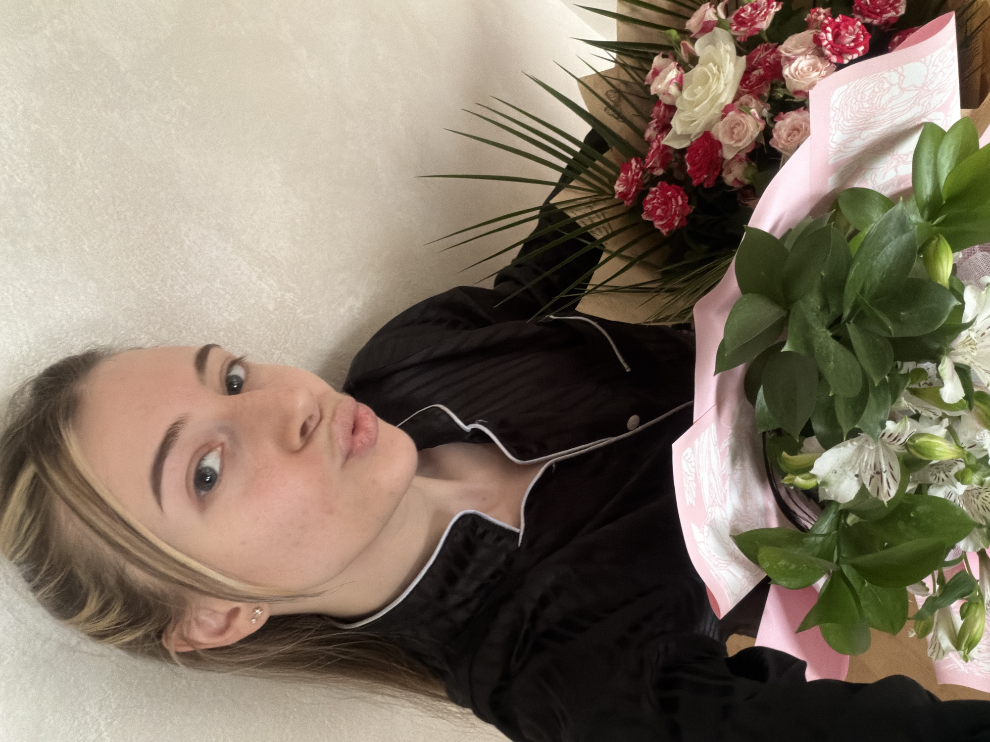 A young woman is captured in a selfie-style shot, lying down indoors with two bouquets of flowers. She appears to be fair-skinned with light brown hair featuring blonde highlights. Her blue eyes are visible, and she is making a playful "duck face" or puckered lips expression. She wears a black long-sleeved pajama top with white piping along the collar and placket. A small, shiny earring is visible in her left ear.
To her right, two distinct flower arrangements are present. The bouquet closer to her head is composed of small red and white flowers, resembling mini roses or spray carnations, intertwined with longer green palm-like foliage. This bouquet is wrapped in natural brown paper. The second bouquet, situated further down, consists of delicate white alstroemeria (Peruvian lilies) with dark green leaves. This arrangement is wrapped in light pink paper, which has a subtle, almost faded, patterned design visible on it.
The background is a plain, light beige or off-white textured wall. The lighting suggests an indoor setting during daytime. The woman's pose and expression indicate a relaxed and perhaps playful mood, likely capturing a moment of receiving or enjoying the flowers. No visible text is present.