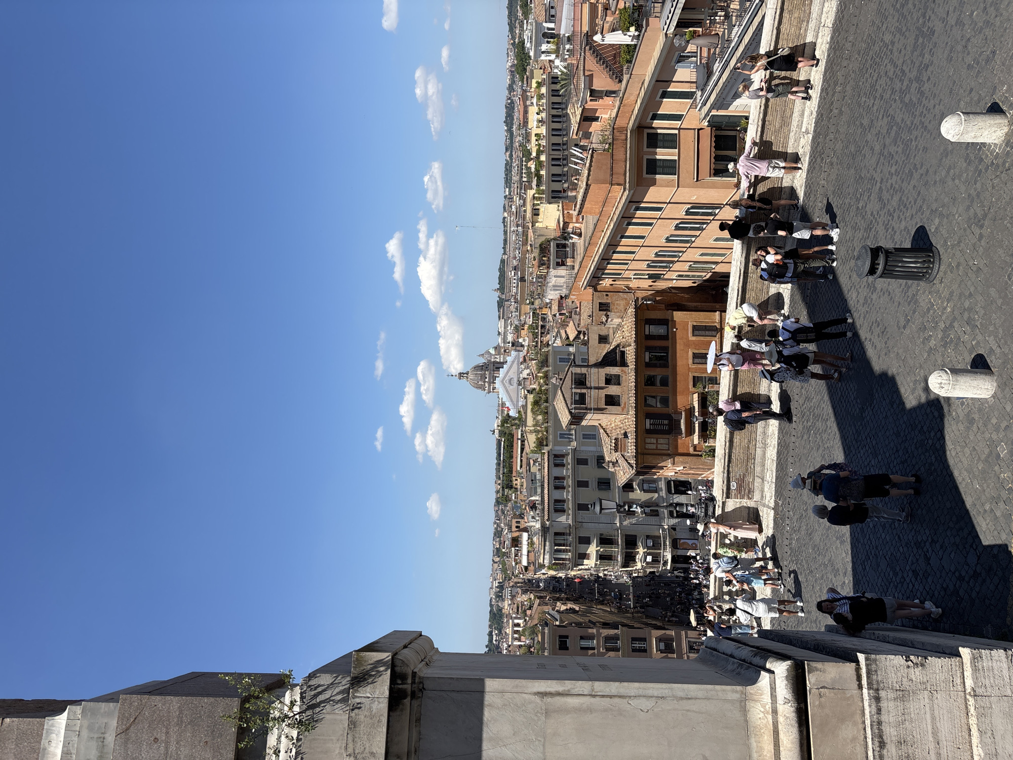 This is a high-angle photograph capturing a street scene in Rome, Italy, likely from an elevated vantage point such as the Spanish Steps, given the architectural style of the buildings and the presence of a grand monument on the left. The sky is a clear blue with a few scattered white clouds. Below, a cobblestone pathway is visible, lined with buildings of various heights and architectural styles, characteristic of Roman cityscape.
Numerous tourists are present on the pathway and on a raised terrace area in the mid-ground. People are seen walking, standing in groups, and taking photographs. Some are dressed in casual summer attire. The pathway is paved with dark cobblestones and has a trash receptacle on the right side. A stone wall separates the pathway from the lower street level, where more buildings and people are visible. The background is filled with a panorama of Roman rooftops and several domed structures, including a prominent dome that resembles St. Peter's Basilica or a similar significant church. The scene is bathed in bright sunlight, casting distinct shadows.