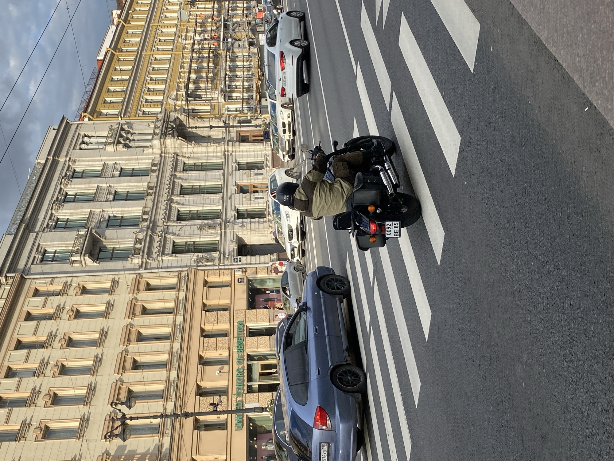 A motorcycle with a rider in a green and tan jacket is crossing a pedestrian crosswalk on a city street in Saint Petersburg, Russia. The motorcycle has a license plate that reads "0092 BE 85". Several cars are on the road behind the motorcycle, and buildings line the street. The buildings are tall and have ornate architecture, typical of Saint Petersburg. The sky is cloudy. One of the buildings has a sign for "UNITED COLORS OF BENETTON". The scene is captured from a slightly elevated, side-view perspective.