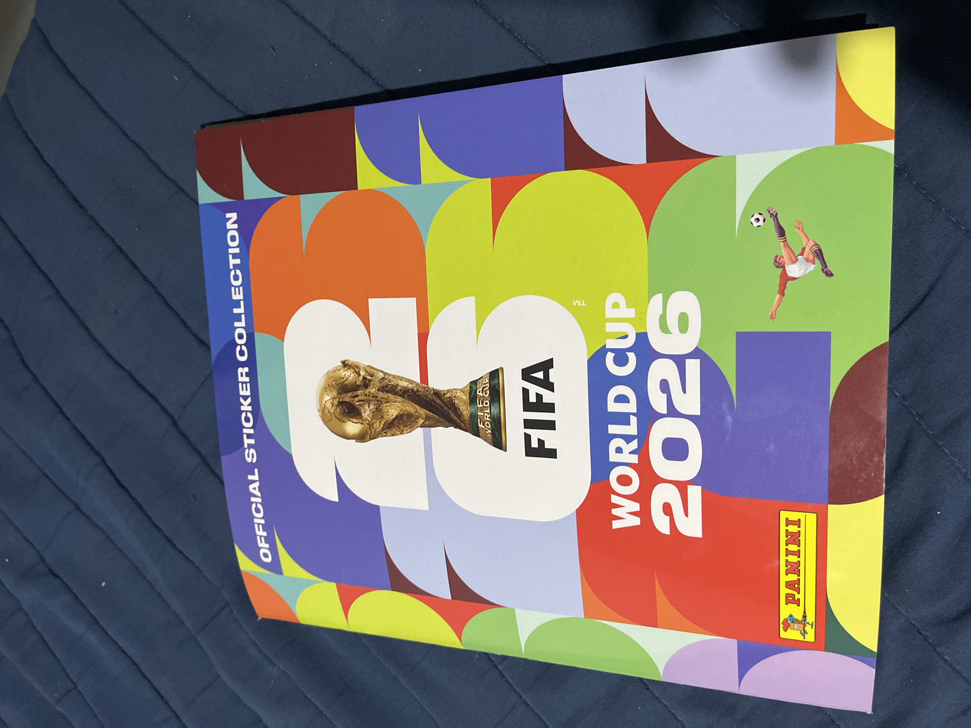 The image shows a Panini sticker collection album for the FIFA World Cup 2026. The album is closed and lying on a blue fabric surface. The cover design features the World Cup trophy, the FIFA logo, and the text "World Cup 2026" in a colorful, stylized design. It also includes the "Panini" logo and the phrase "Official Sticker Collection". There are no people visible in the image. The location is not apparent.