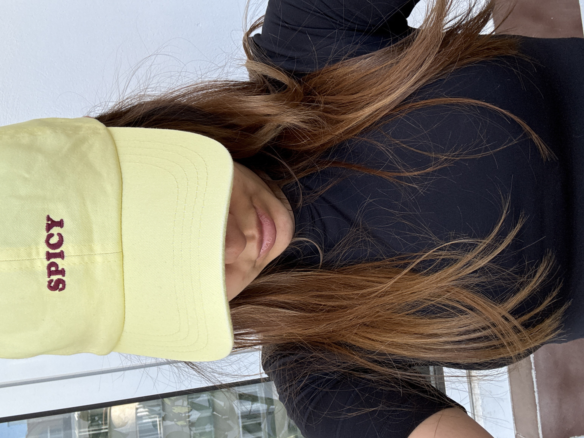 The image shows a woman wearing a black top and a yellow baseball cap with the word "SPICY" embroidered in red. Her long, light brown hair is visible around her face and shoulders. The background includes a window with a view of buildings, suggesting an urban setting. The location is possibly Sofia, Bulgaria. The perspective is from slightly above and to the side of the subject.