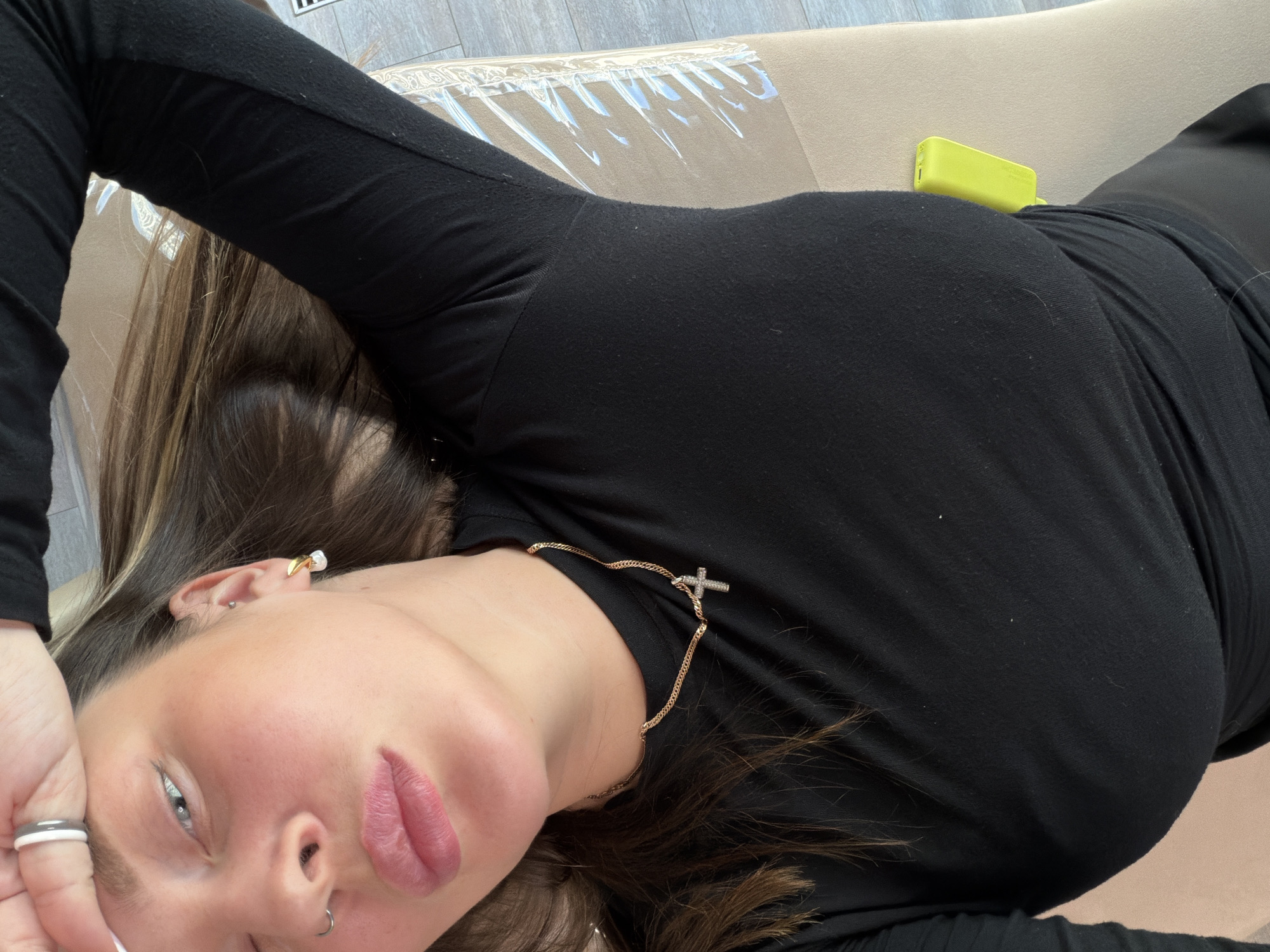 A woman with fair skin and light blue eyes is lying down on a beige upholstered surface, possibly a couch or bed. She is wearing a black long-sleeved shirt and a gold necklace with a cross pendant. She has brown hair and is wearing rings on her fingers. Her left hand is partially covering her forehead. A small yellow object is visible near her waist. The background consists of a light-colored floor. The location in the city of Dolgoprudny, Russia is not discernible from the image.
