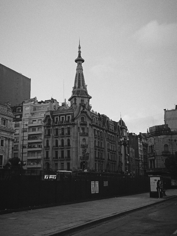 This black and white photograph captures an urban street scene in Buenos Aires, Argentina, dominated by the Palacio de la Prensa (Casa de la Cultura) at Avenida de Mayo 575, a prominent building known for its ornate Beaux-Arts architecture and distinctive spire.
In the foreground, a dark, solid construction barrier or hoarding runs horizontally across the lower half of the image, obscuring the ground floor of the buildings. Several indistinct posters are affixed to this barrier. A paved street with a curb delineates the lower right corner, leading to a sidewalk on the right.
To the far right, two individuals are discernible standing next to a bus stop shelter. One person is mostly obscured by the shelter, while the other stands adjacent, facing towards the street. Their actions are static.
The midground is dominated by a row of multi-story buildings. The Palacio de la Prensa occupies the central frame, distinguished by its intricate detailing, numerous balconies, dormer windows, and a multi-tiered cupola culminating in a tall, slender spire. Its facade shows multiple floors with arched windows. To its left and right, other multi-story buildings, some with balconies and classical architectural elements, line the street. One building to the far left is a stark, unadorned structure, contrasting with the classical styles.
The sky above is uniformly light grey, indicating an overcast day. The overall environment is an established urban setting with mixed architectural styles, captured under diffuse natural light. No active vehicles or clear interactions between people are visible.