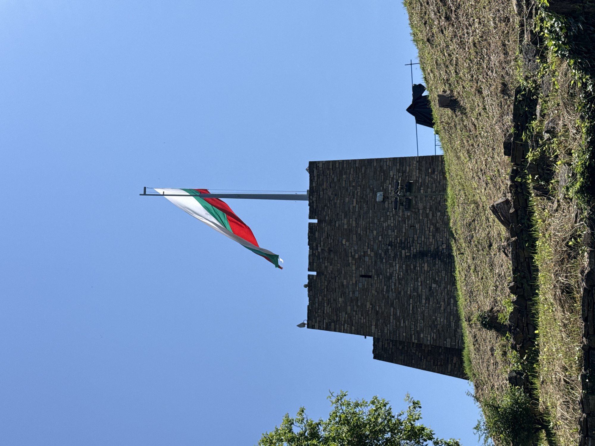 The image depicts a low-angle shot of a flag waving in the wind in Veliko Tarnovo, Bulgaria. The flag, with white, green, and red stripes, is mounted on a pole extending from a stone structure with crenellations. Part of the structure is covered in greenery and moss. The sky is clear blue. Tree tops are visible at the bottom of the image.