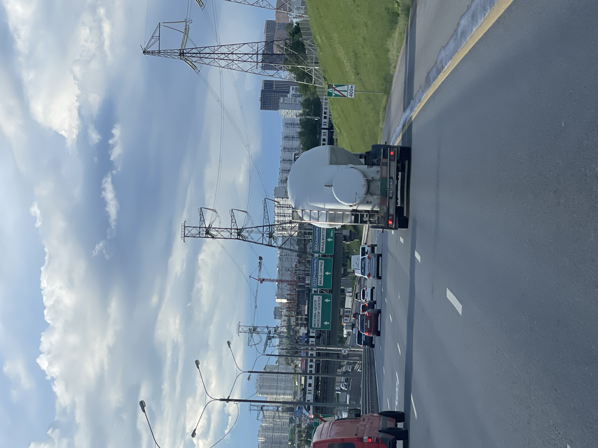 The image shows a multi-lane highway in Novodrozhzhino, Russia. Traffic is present, including a tanker truck. Overhead power lines are visible against a partially cloudy sky. Road signs indicate directions to Moscow-Center, A101 Roslavl, MKAD-West, M4 Kashira, and MKAD-East. A sign also indicates a distance of 400 meters. Buildings, including apartment complexes and construction sites with cranes, are in the background. Streetlights line the side of the highway.