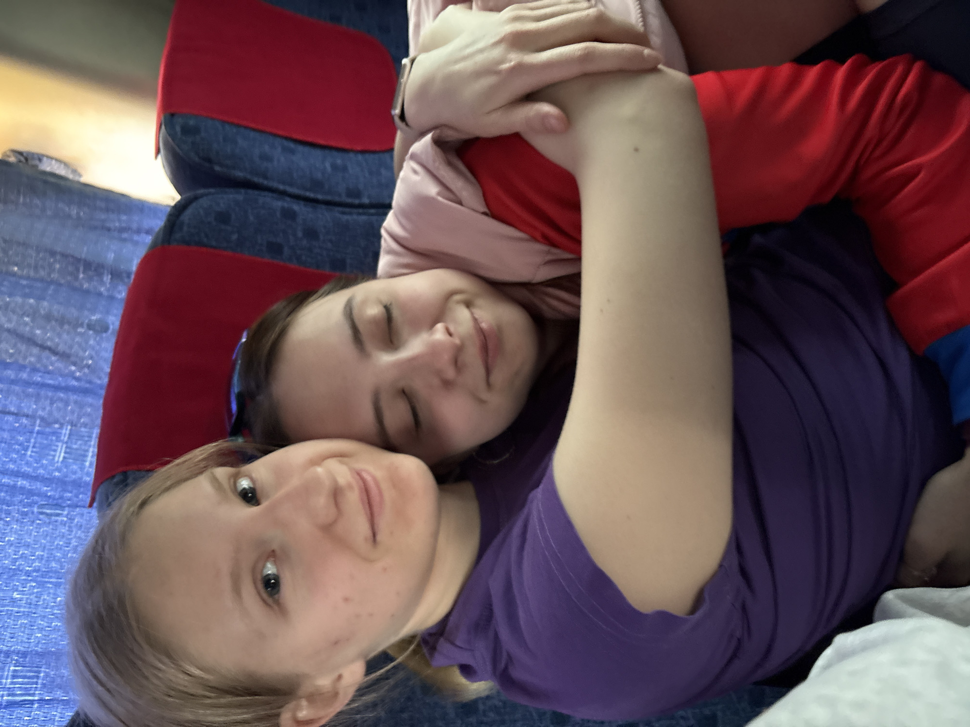 Two young women are embracing on a blue and red patterned surface. One woman, with blonde hair and freckles, is facing the camera. The other woman, with brown hair, has her eyes closed and is wearing a pink jacket. The blonde woman is wearing a purple shirt, and the brown-haired woman is wearing a red shirt. The location is unidentifiable as Verkhnyaya Dobryanka, Russia.