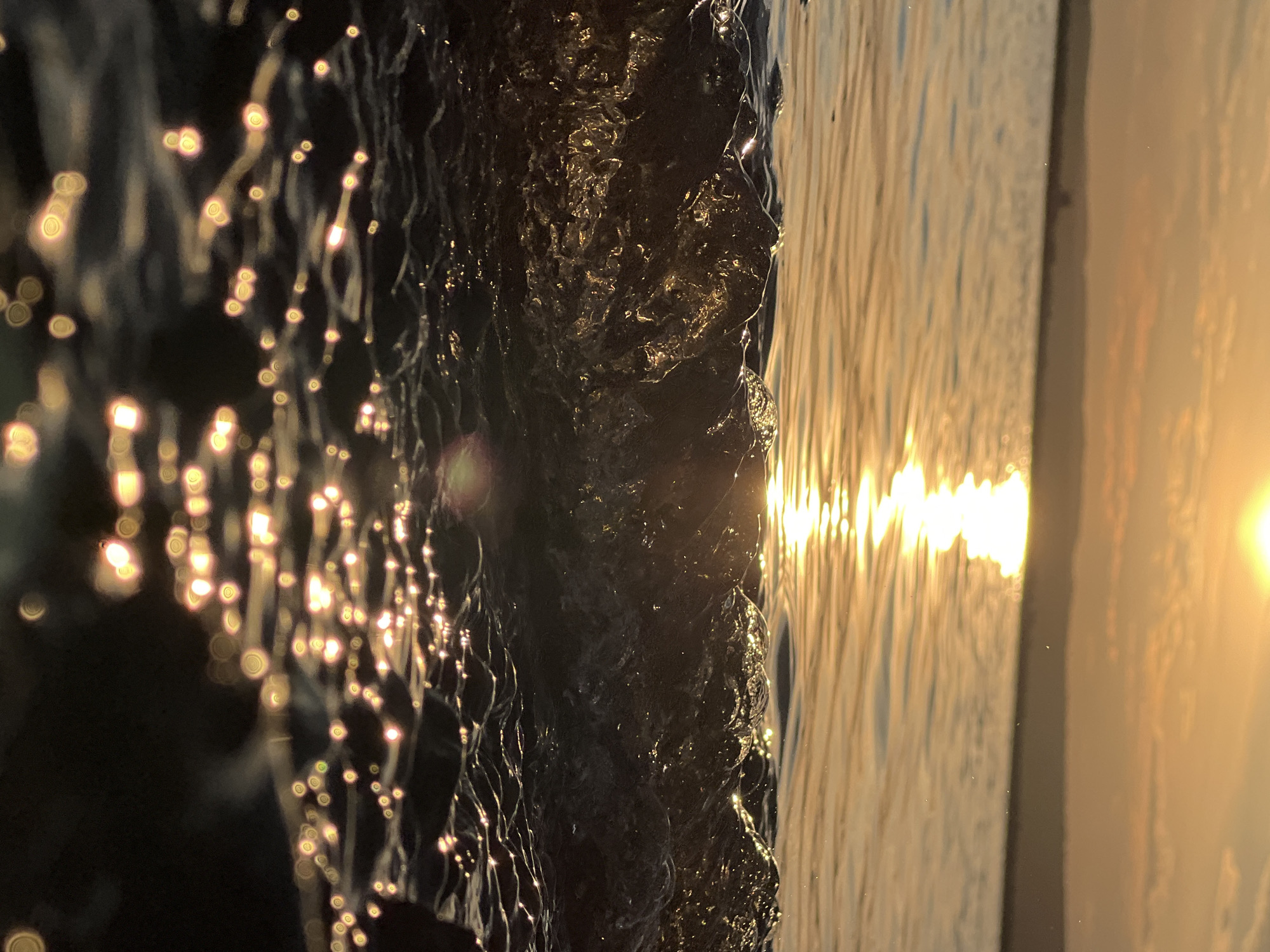 This media file captures a close-up, vertical view of water flowing or cascading, likely part of a fountain or waterfall, set against a dark background. The water's surface is dramatically illuminated by what appears to be the setting sun. The sun's rays are reflected in the water, creating a shimmering effect with bright, bokeh-like highlights. The foreground water appears dark and textured, with individual droplets or streams catching the light. The background on the right side shows a smooth, golden-hued surface, also reflecting the sun's light, possibly indicating the water is falling onto another body of water or a reflective surface. The overall mood is tranquil and luminous, emphasizing the interplay of light and water. The time of day is clearly sunset, given the warm, golden light and the low angle of the sun. There are no people or man-made objects explicitly visible, focusing solely on the natural elements and light. The location is identified as Chaikovsky, Russia.