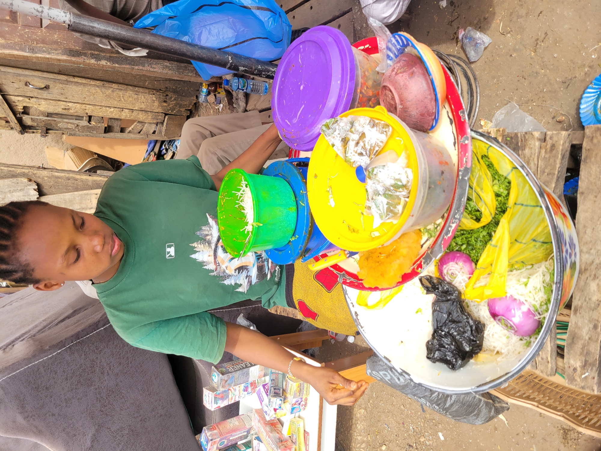 This image captures a vibrant street food stall in Jos, Nigeria, during daylight hours. The primary subject is a person, likely a vendor, wearing a green t-shirt with a white Major League Baseball (MLB) logo. Their hair is styled in braids, and their eyes are cast downwards as they reach their right hand towards a lower shelf holding various small cardboard boxes. One box clearly displays the word "CLEANER," and another shows "QUICK ACTION."
The vendor operates from a makeshift stall featuring numerous colorful plastic containers holding food items. Prominent are a large purple lid, a bright yellow container with clear plastic bags of what appears to be prepared food, and a green container filled with shredded light-colored vegetables or noodles. A large, round, colorful serving bowl or tray is central to the setup, filled with shredded white ingredients, chopped green herbs, sliced red onions, a dark, wrapped item (possibly meat or another food item in a black bag), and a yellow plastic bag. Another smaller, lighter-colored fried item (possibly a snack like akara) is visible on a plate.
In the background, another person's legs are partially visible, wearing light-colored trousers. The stall is constructed from rough wooden planks and crates, with a black metal pipe running across the upper part of the frame. The ground is a mix of dirt and possibly concrete, with scattered debris including empty plastic bottles and discarded plastic bags. A large blue plastic bag rests near the wooden structures. The scene suggests a busy market or roadside environment where food and possibly other small goods are sold.