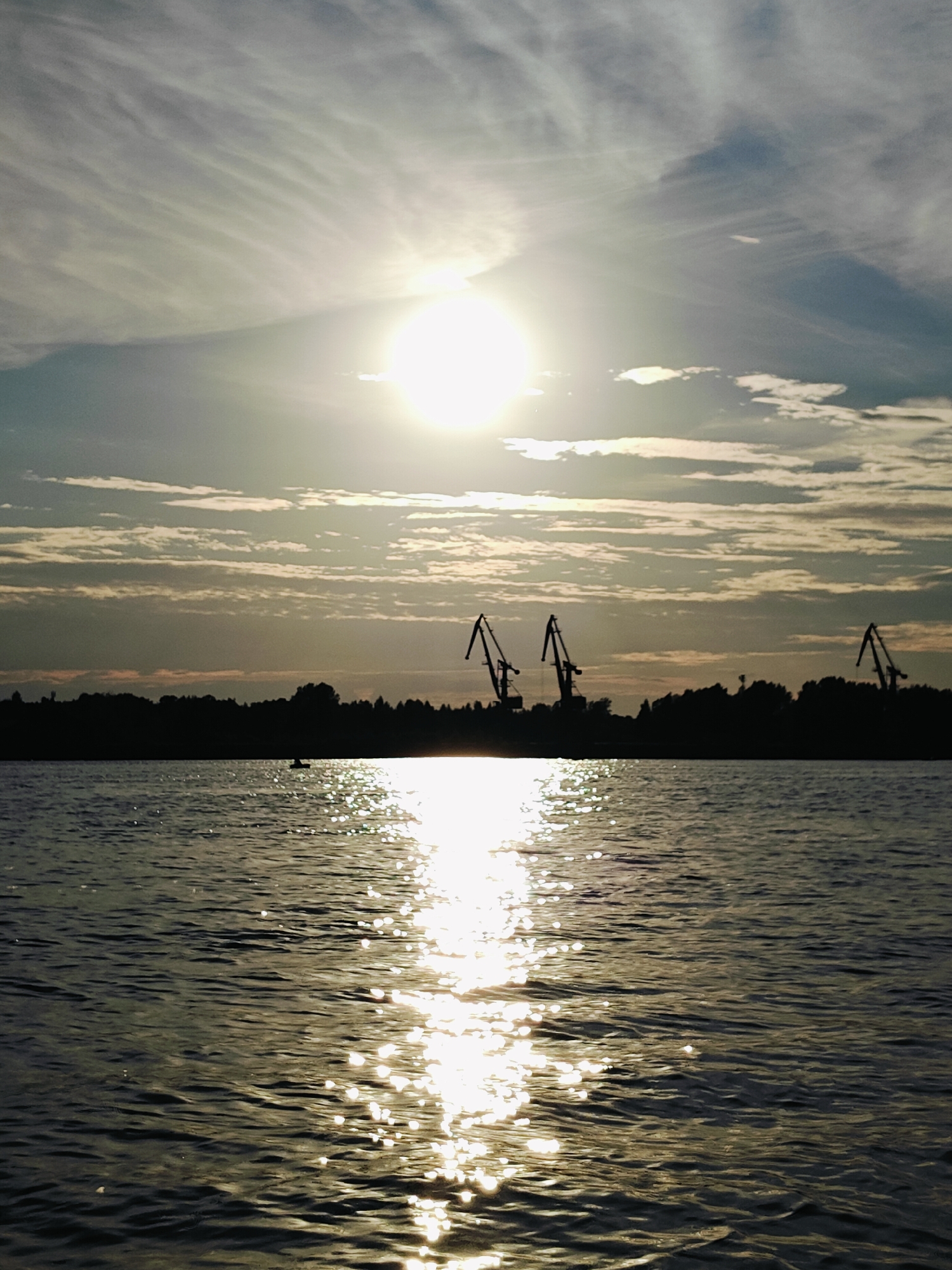 A wide shot captures a sunset over the Kama River in Chaikovsky, Russia. The sun, a bright, hazy orb, is positioned in the upper center of the frame, partially obscured by wispy clouds. Its light reflects off the rippling water in a shimmering path that extends towards the viewer. In the distance, a dark silhouette of trees lines the riverbank. Three industrial cranes are visible against the horizon, their skeletal structures stark against the twilight sky. A small, dark silhouette of a boat with a person in it is on the water, in front of the tree line. The sky is a mix of pale blue and soft grays, with elongated clouds stretching horizontally across the frame. The overall mood is serene and tranquil.