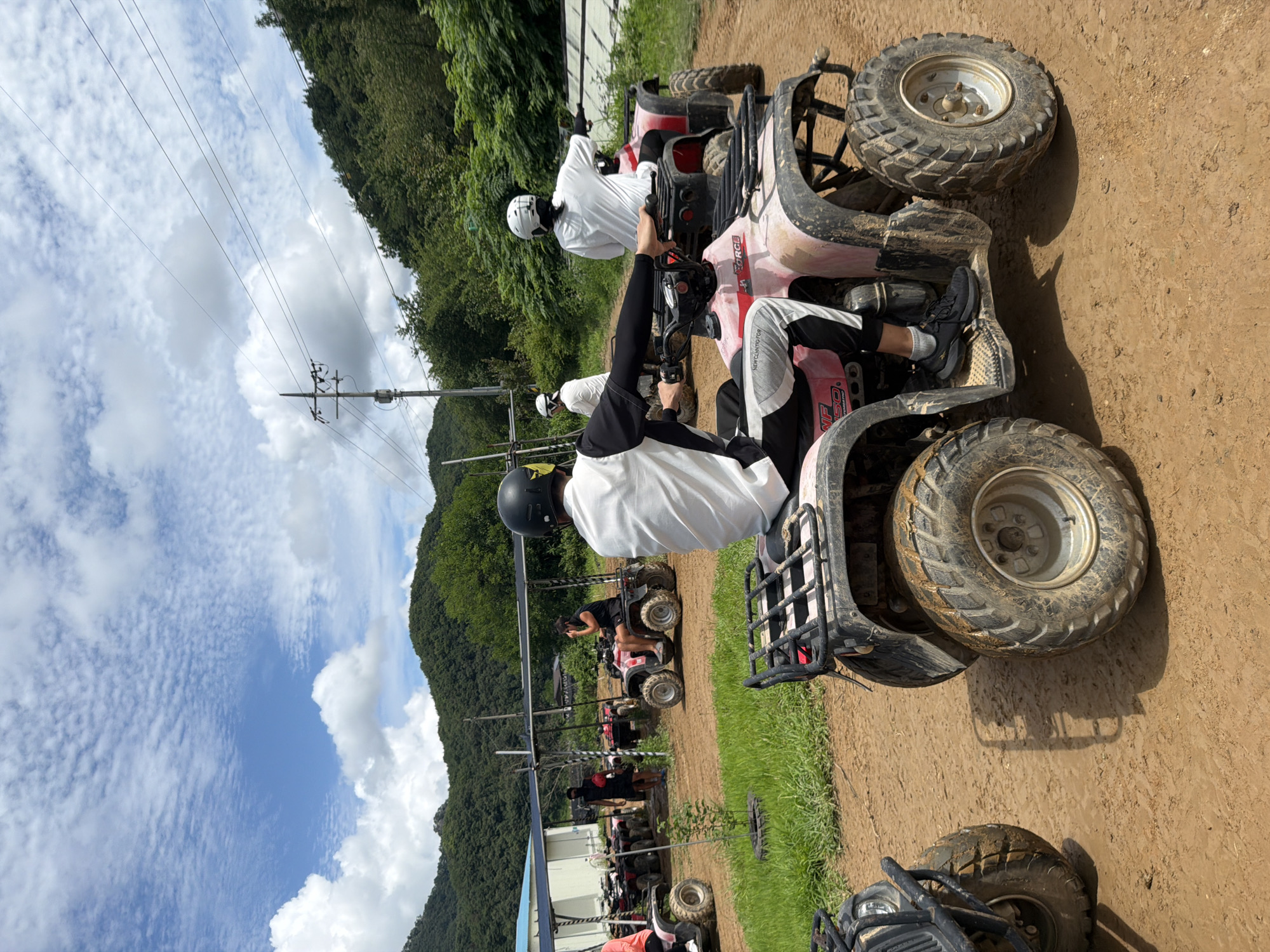 In Seo-myeon, South Korea, several individuals are engaged in an ATV (All-Terrain Vehicle) riding activity. The scene is set on a dirt track, with lush green hills and trees forming the backdrop under a partly cloudy sky. In the foreground, a pink ATV is prominently featured, with a person wearing a black helmet, a white long-sleeved shirt, and black pants with white stripes seated on it. This person is holding onto the handlebars, appearing to be ready to ride or in the process of riding.
Behind this ATV, another ATV is visible, driven by a person in a white shirt and helmet. Further in the background, more ATVs are parked or being ridden, and several other individuals are present. Some are on ATVs, while others are standing around, possibly as instructors or waiting for their turn. A covered structure, likely a shelter or waiting area, is visible on the left side of the image, along with parked ATVs. The ground is a mix of dirt and some muddy patches, indicating the terrain for the activity. The overall environment suggests an outdoor recreational area designed for ATV riding.
