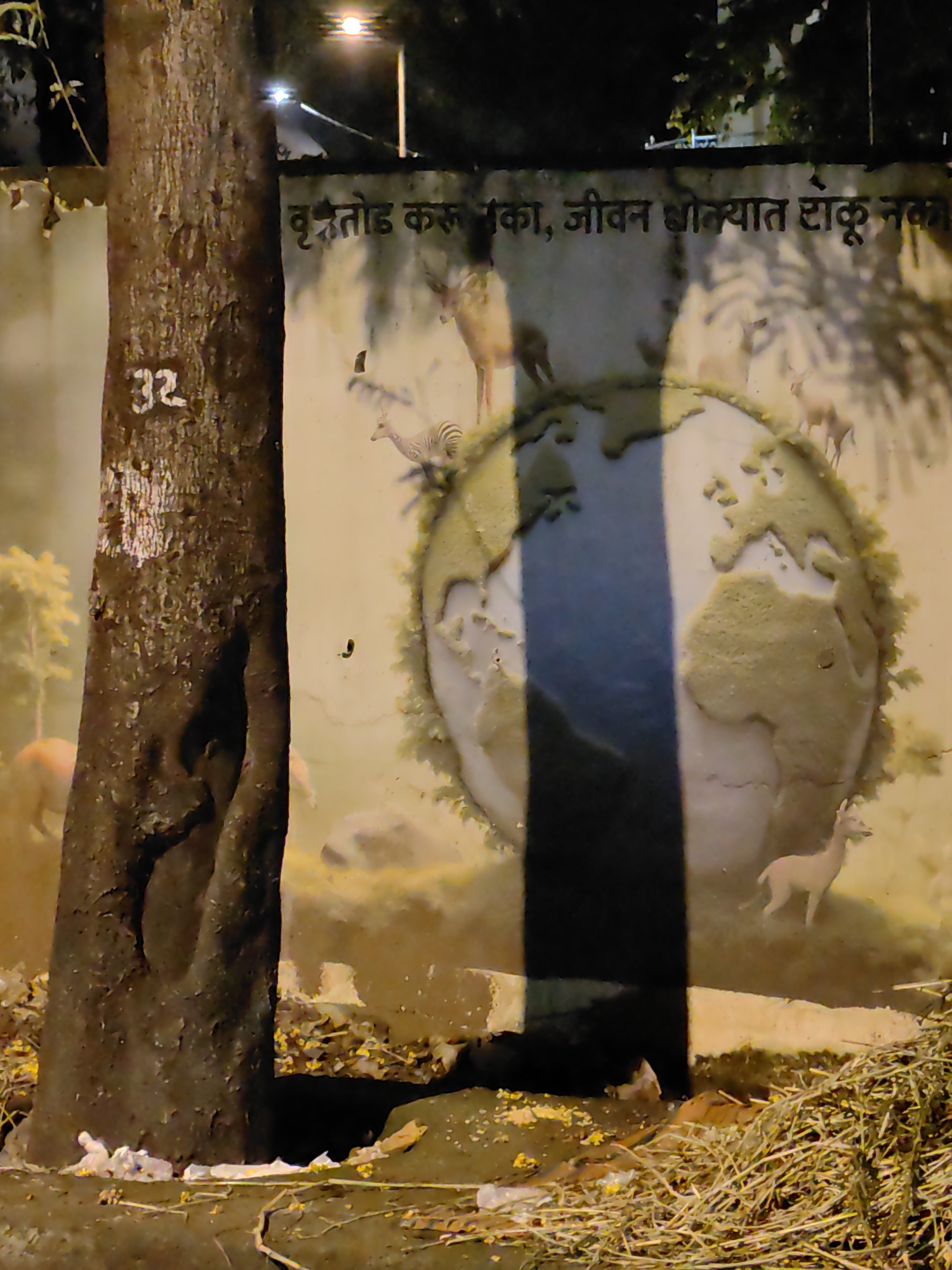 The image depicts a mural painted on a wall in Mumbai, India. The mural features a globe with various animals, including deer and a zebra, surrounding it. A large tree trunk stands in the foreground, obscuring part of the mural. The text on the mural is in Marathi and appears to be an environmental message, possibly related to trees and life. The scene is illuminated by artificial light, casting shadows. The ground is covered with debris, including dry leaves and twigs. A number "32" is painted on the tree trunk.