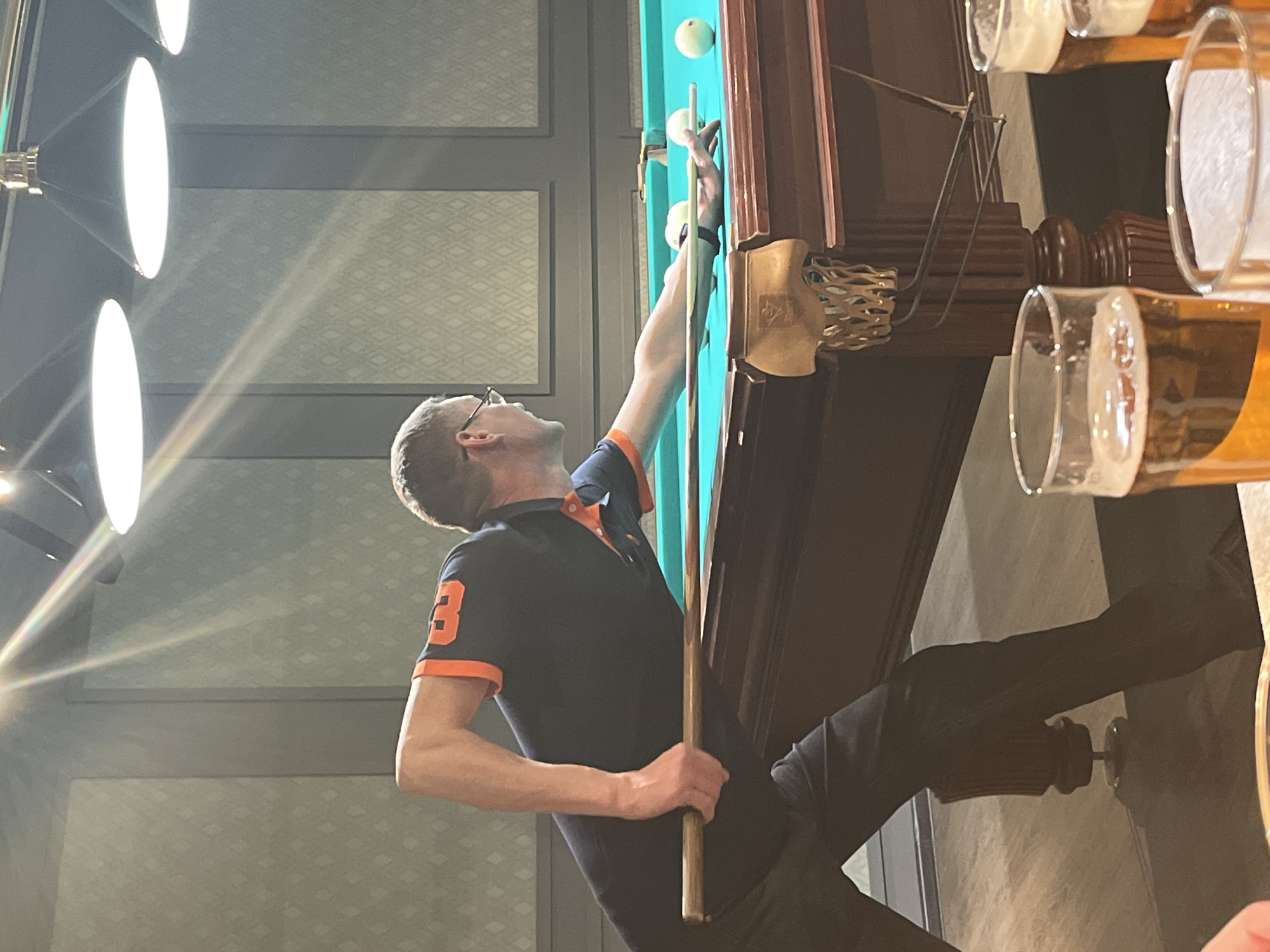 A man with short hair and glasses, wearing a dark polo shirt with orange trim and the number "3" on the sleeve, is playing pool at a table. He is extending his arm towards the table with a pool cue in his hand. The pool table is a light teal color with white pool balls visible. To the right of the pool table, there are partially consumed glasses of beer. The setting appears to be a billiards hall in Izhevsk, Russia, characterized by interior design details such as lighting fixtures and wall patterns. The man is focused on aligning his shot.