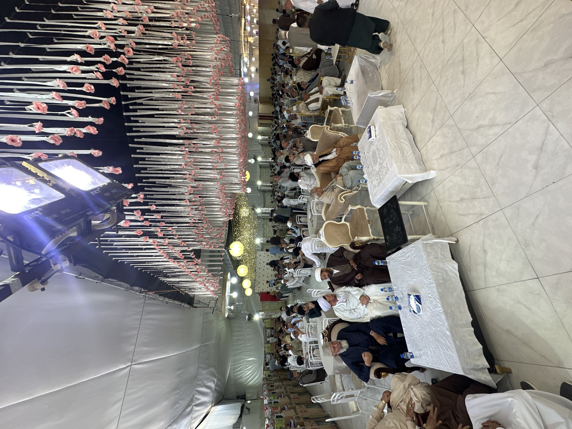 Here is a description of the image:
The image depicts a large gathering of people seated at tables in what appears to be a banquet hall or event space in the city of Quetta City Tehsil, Pakistan. The attendees are predominantly men, many wearing traditional Muslim attire, including head coverings and robes. They are seated around tables draped with white tablecloths, each containing water bottles and tissue boxes. The tables are set in rows, suggesting a formal setting, possibly a wedding. The hall is decorated with long strips that have roses on them on the ceiling. There are also stage lights on the ceiling. The floor has white tiling.