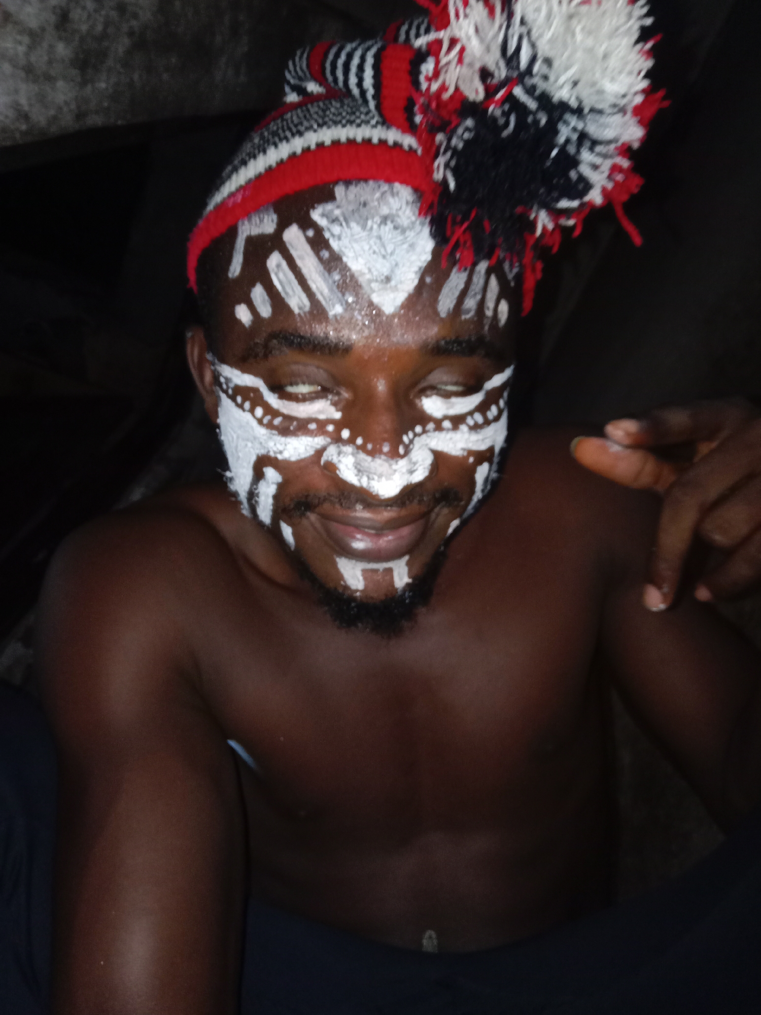 A dark-skinned man with white face paint is looking down and smiling. He is wearing a striped hat with a pom-pom on top. He is shirtless. A hand is partially visible in the upper right. The environment appears to be dark and undefined. The location and city are unknown.