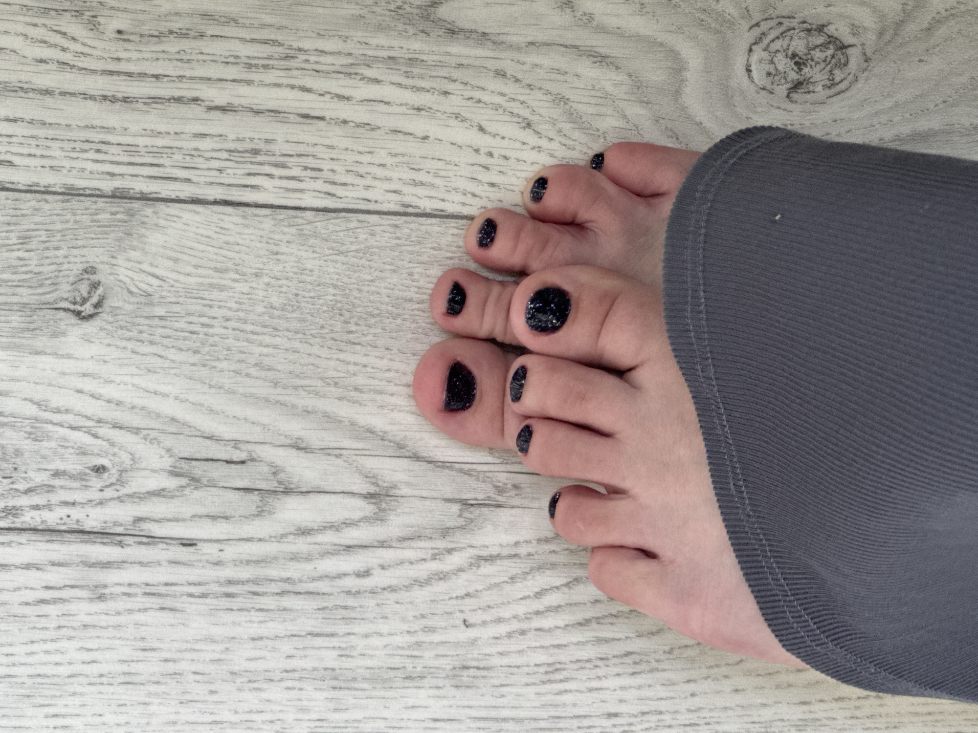 This media file features a close-up view of a person's feet, which are resting on a light-colored wooden surface. The feet are adorned with dark, glittery nail polish on each toe. A portion of a dark gray, ribbed fabric garment is visible, partially covering the person's leg. The wooden surface has a visible grain pattern with some knot details, suggesting a rustic or natural setting. The lighting appears to be even, with no strong shadows, indicating it could be daytime indoors or outdoors with diffused light. There is no visible text in the image. The focus is on the details of the feet and the texture of the surfaces. The overall impression is one of personal care and a simple, possibly domestic, environment, given the mention of a rural council in Belarus.