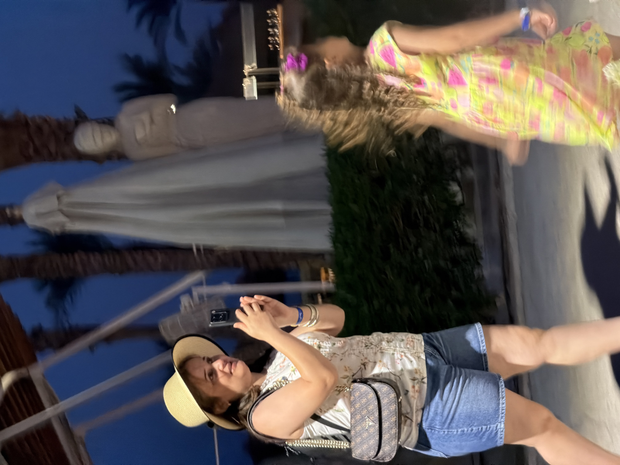An outdoor scene at dusk in Sharm El Sheikh, Egypt, shows a woman in a straw hat and floral print top taking a photo with her phone. She is wearing denim shorts and a crossbody bag with a monogram pattern. To her right, a young girl in a brightly colored dress with a pink bow in her hair is blurred due to motion. In the background, palm trees and indistinct statues are visible against a dark blue sky. The lighting suggests artificial illumination from streetlights or buildings.
