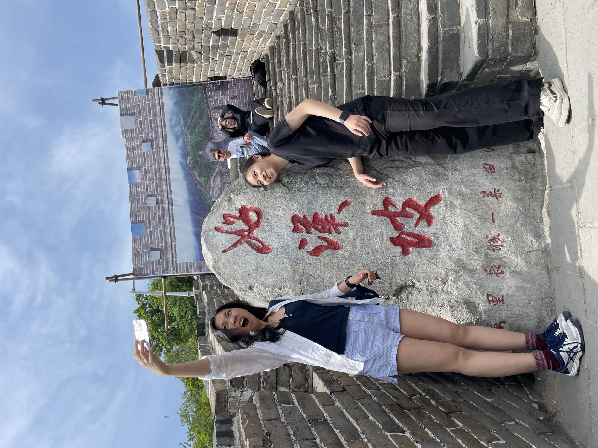 The image shows two young women lying on a stone at a tourist location, probably the Great Wall of China. The woman in the foreground holds up a smartphone, taking a selfie. She wears a dark blue shirt, light blue shorts, a white shirt, and sneakers. Her mouth is open in a joyful expression. The woman in the background is wearing a black shirt, black pants, and white sneakers. She smiles toward the camera. Behind them is a sign with Chinese characters, and behind that sign is a picture of a section of the Great Wall. The Great Wall itself forms the background. The city is Huairou District, China.
