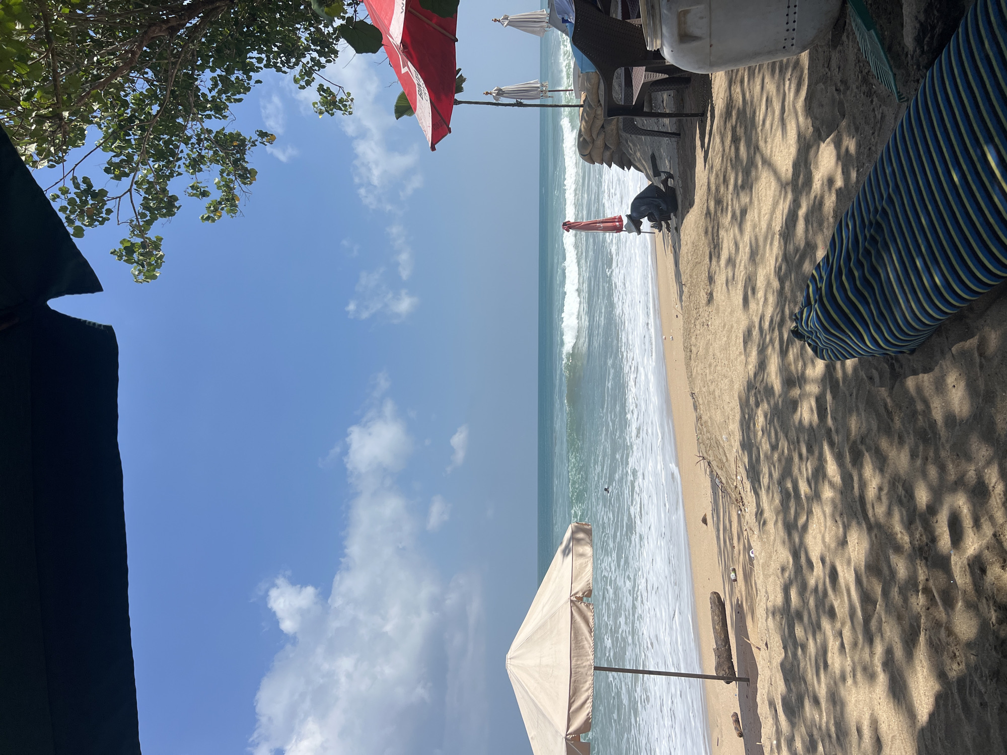 A sunny day at Kuta Beach, Indonesia, is depicted from a low angle, possibly from a beach chair or lounger. The foreground is dominated by sand with dappled shadows from overhead foliage, suggesting a tree nearby. A blue and yellow striped fabric object, perhaps a cushion or rolled-up mat, lies on the sand. To the right, a large white plastic container and a green rake are visible, along with dark chairs and a red beach umbrella. Sandbags are stacked behind the chairs. In the mid-ground, the sandy beach slopes towards the ocean, with a beige beach umbrella on the left and another red umbrella and two white, closed umbrellas further down. A person wearing a hat and dark clothing is crouched near the water's edge, appearing to work on something. The ocean displays moderate waves with white foam crashing onto the shore. The sky is bright blue with scattered white clouds. The overall scene captures a typical beach day setting with elements of relaxation and activity.