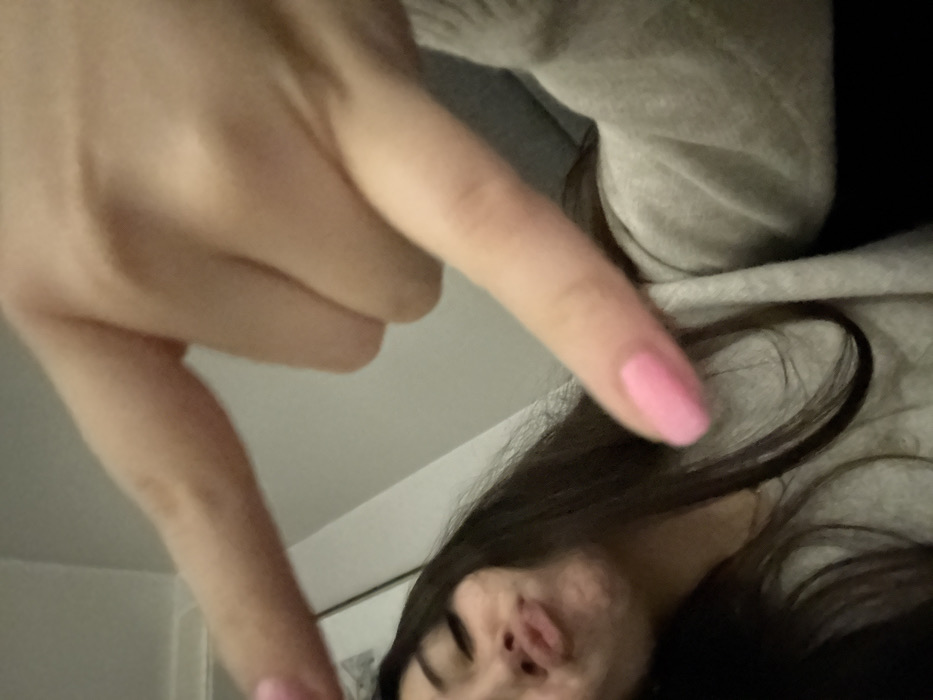 A person is taking a selfie with their hand gesturing in the frame, with pink nail polish. The subject has long, dark hair and is wearing a light-colored sweatshirt. The background is mostly out of focus and appears to be a white wall. The focus is on the hand and the person's face. The angle is slightly from above, giving a distorted perspective.