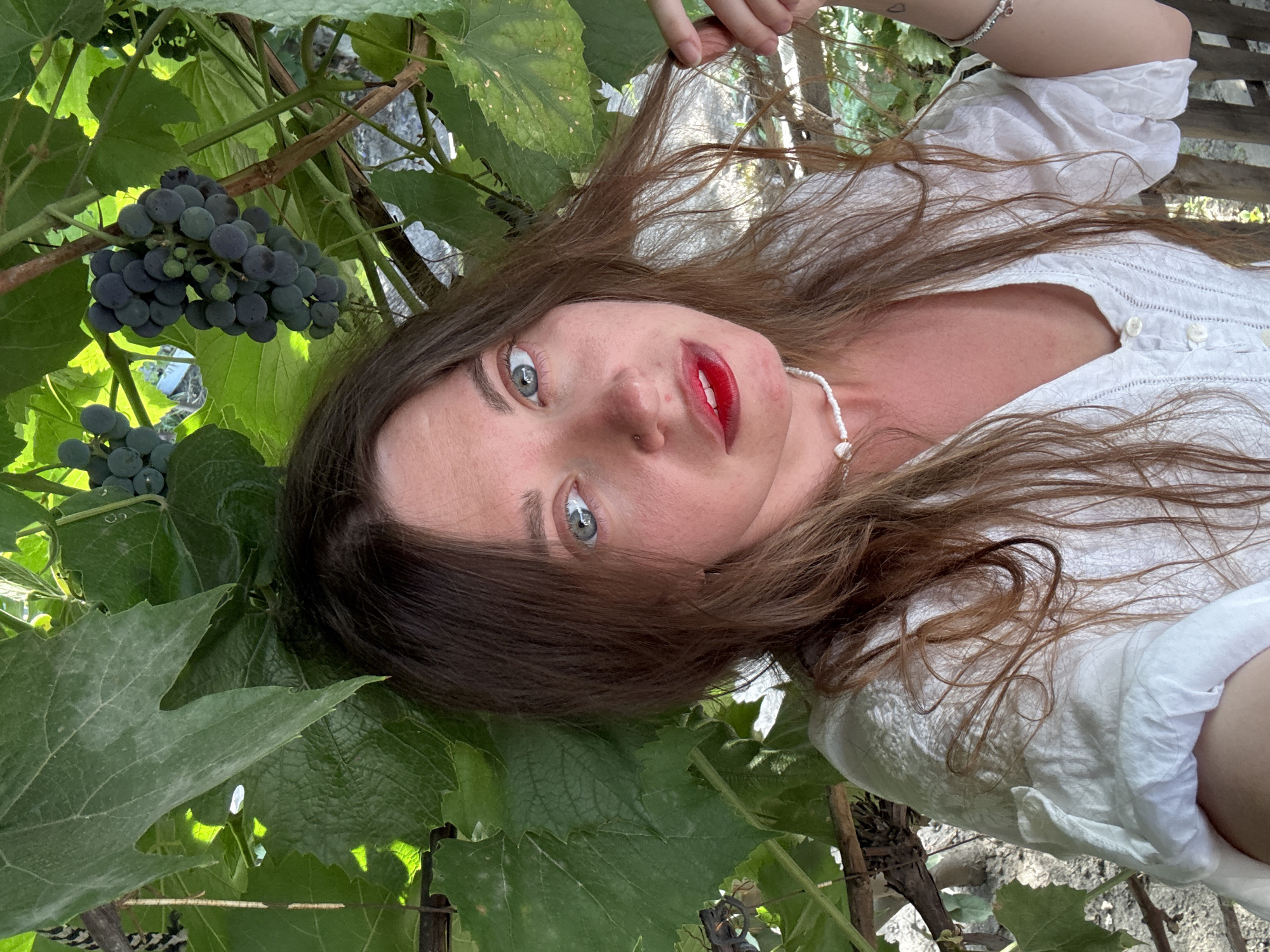 A young woman with long, brown hair and blue eyes is pictured from the chest up, framed by green grape leaves and dark grapes. She is wearing a white, button-down shirt and a pearl necklace. Her lips are painted red. The background shows a rustic wooden fence and hints of foliage, suggesting an outdoor setting in a rural or garden area. The image was taken in the city of Sol-Iletsk, Russia.