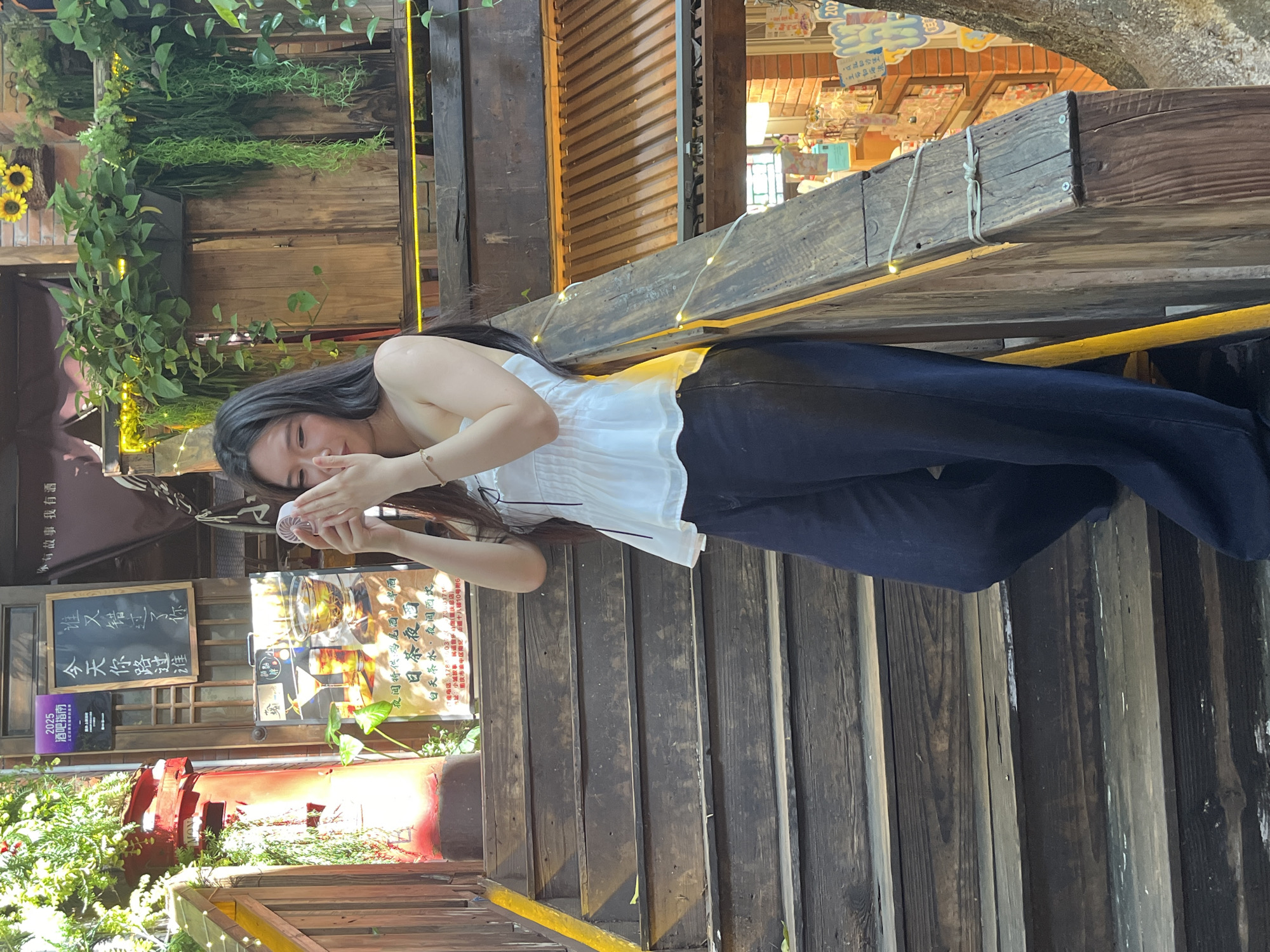 A young woman with long, dark hair stands on a wooden staircase, holding a small white fan to her face. She is wearing a white strapless top and dark wide-leg pants. The staircase is outdoors, with wooden railings and steps. Various plants hang from above, and there are signs and decorations on the wall behind her. The overall scene appears to be in an urban, possibly commercial or tourist area. The woman's expression is neutral as she holds the fan. The lighting suggests it is daytime.