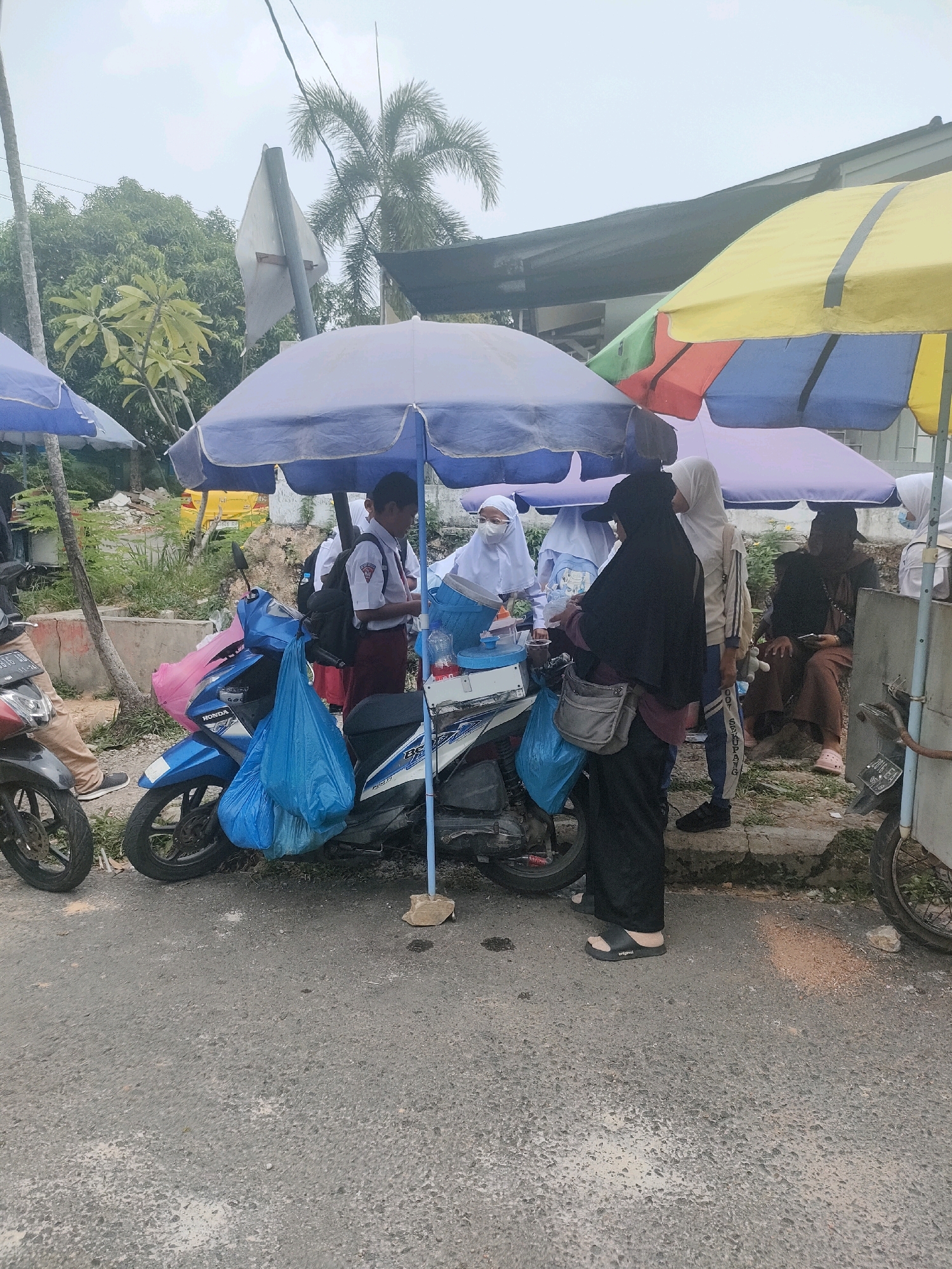 The image depicts a street scene in Batam, Indonesia, under an overcast sky, with several people gathered around a temporary stall set up on a motorcycle. The stall is covered by blue and purple umbrellas. A young boy in a school uniform, identified as Indonesian by his attire, is standing at the stall, possibly purchasing something. Two other individuals wearing hijabs are also present at the stall. To the right, another person is seated and appears to be a customer. A red motorcycle is parked on the left, and a blue motorcycle, likely part of the stall setup, is in the center. Blue and pink plastic bags are hanging from the motorcycle. The environment appears to be a roadside setting with some vegetation and debris in the background. The ground is paved with asphalt.