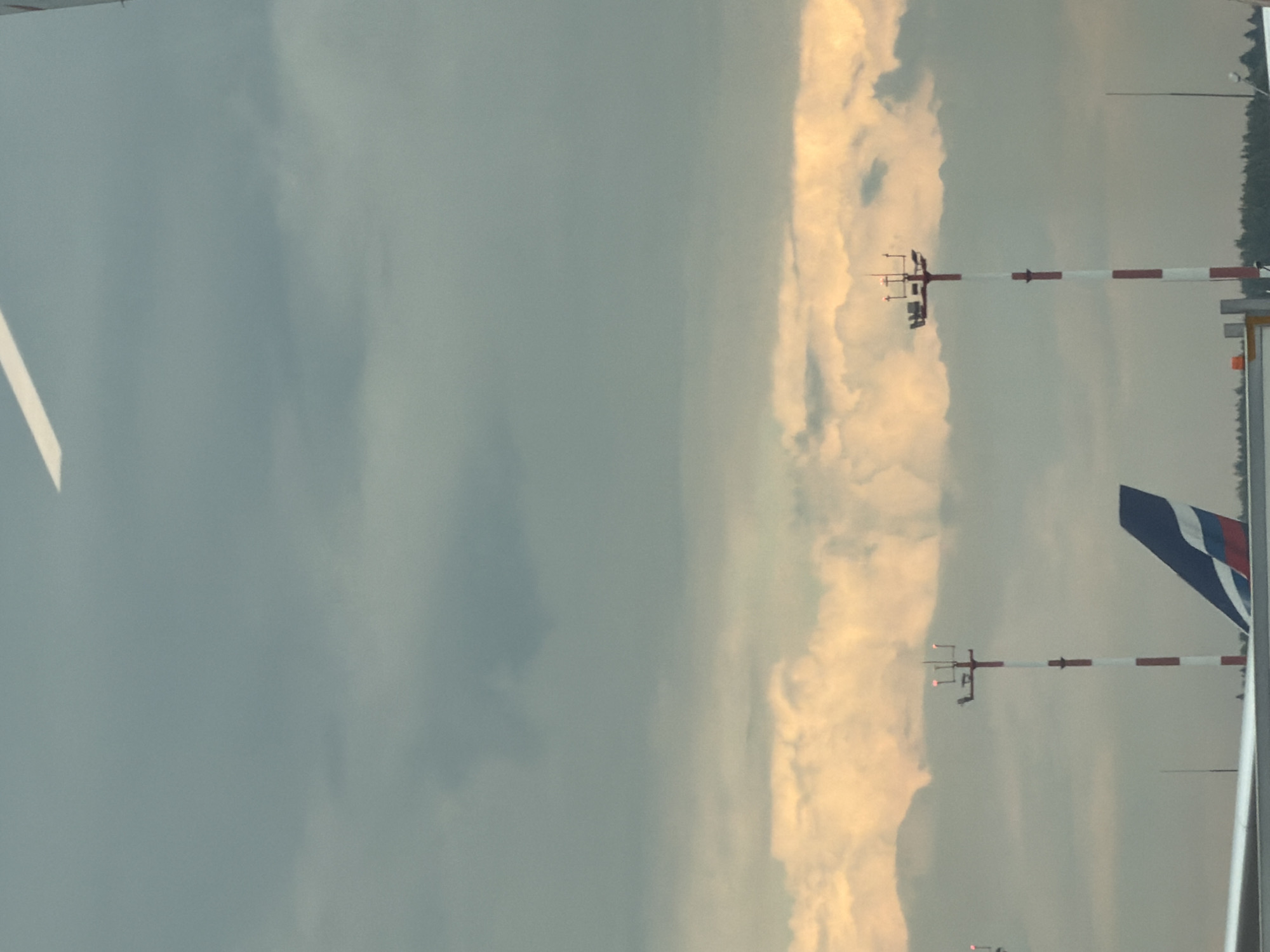 The image depicts an airport scene in Khimki, Russia. The sky is overcast with some illuminated clouds suggesting either dawn or dusk. In the foreground, the wing of an aircraft is visible, and further back, the tail fin of another aircraft with a red, white, and blue design is prominent. Two tall, red and white striped towers, likely navigation or communication aids, stand on the tarmac, each topped with lights. The background shows a treeline, indicating the presence of vegetation beyond the airport grounds. The overall impression is one of an operational airport under a moody sky.