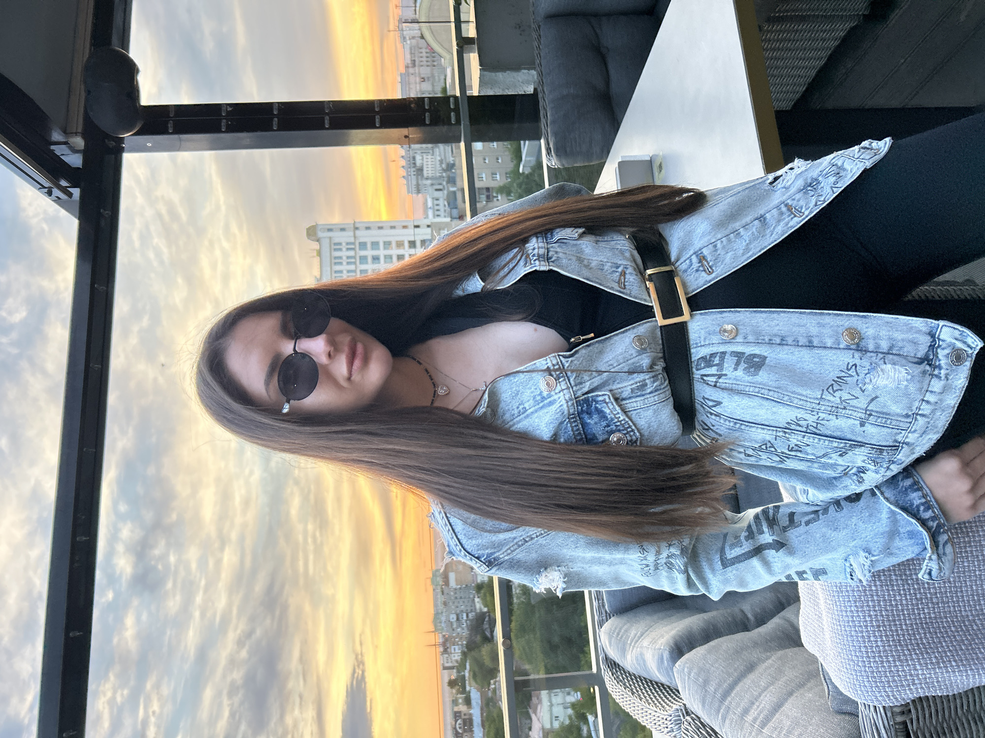 A young woman with long, dark hair and sunglasses is seated on a balcony overlooking a cityscape during sunset. She is wearing a denim jacket over a black top and black pants. The jacket is adorned with graffiti-like text and has distressed detailing on the cuffs and hem. A black belt with a gold buckle cinches her waist. She is wearing a necklace with a pendant. The sky is filled with dramatic clouds painted in hues of orange, yellow, and purple, indicative of a beautiful sunset. The background shows buildings and trees of a city, suggesting an urban setting. The overall mood is relaxed and contemplative, with the woman seemingly enjoying the view. The time of day is clearly sunset.