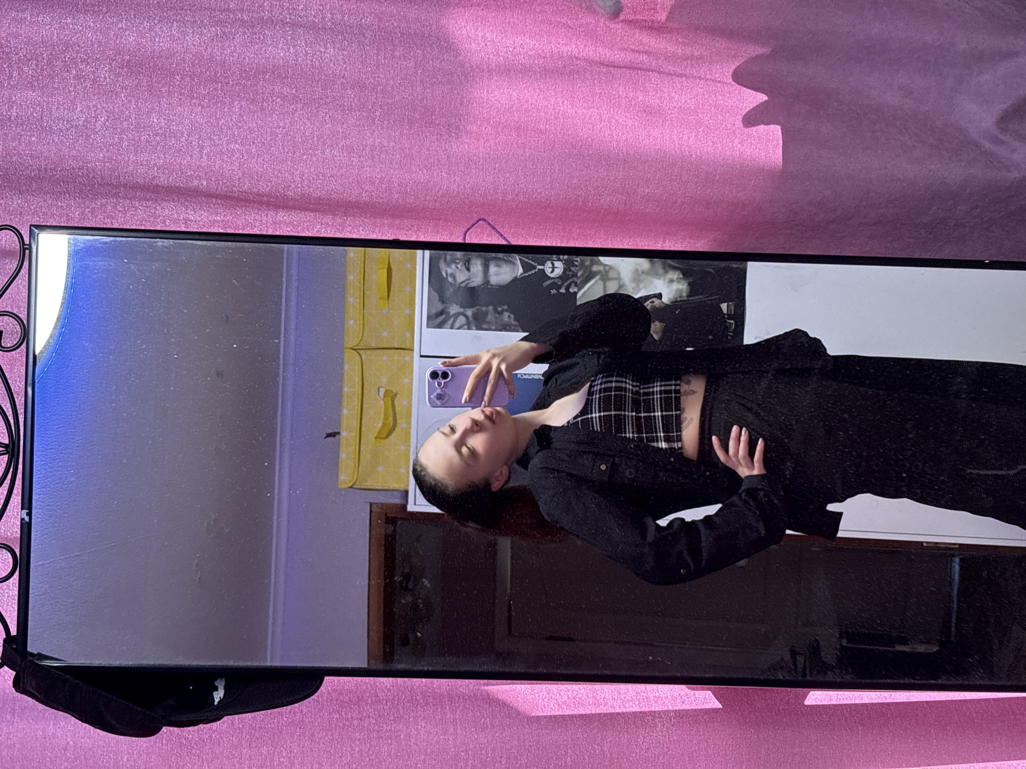 A mirror reflects a young adult taking a selfie. The person is wearing a black jacket over a plaid top, and black pants. Their phone is purple. Behind them is a poster featuring a black and white image of a person, as well as a yellow bag. The mirror is resting on a pink surface. The location is Прикамский, Russia.