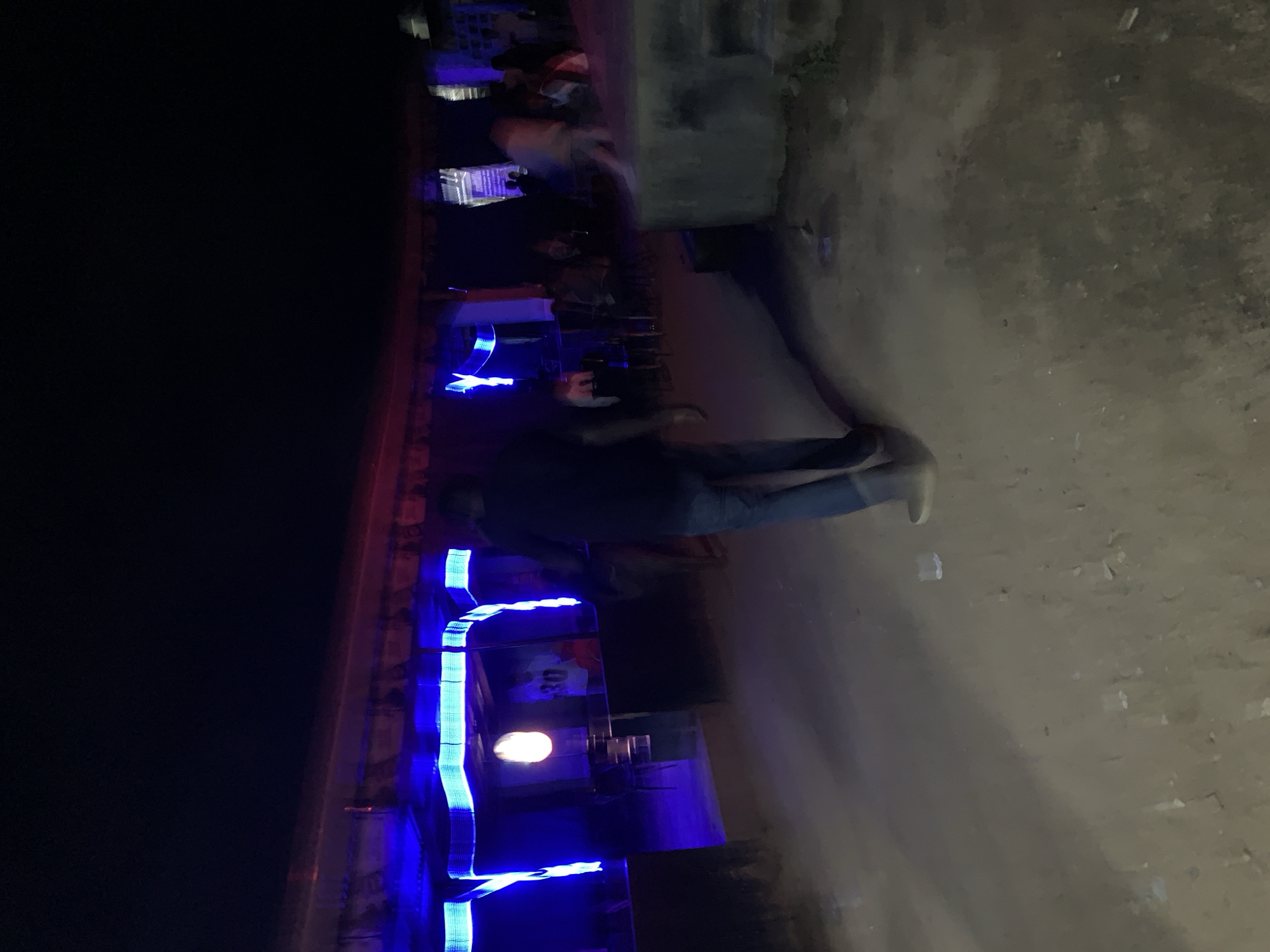 The image depicts a nighttime scene at an outdoor social gathering or informal establishment, likely a bar or lounge, located in the New Stadium area of Nigeria.
The foreground features an unpaved ground of dirt or sand, scattered with small, light-colored debris. A person, dressed in dark trousers and a dark top, walks away from the viewer towards the background structure, exhibiting motion blur on their lower body. To the right, a large, rough concrete block or low wall is visible, with another heavily blurred figure moving past it.
In the midground and background, a single-story structure spans the scene, illuminated by bright blue neon strip lighting that outlines its low roofline and vertical supports. A subtle reddish hue emanates from above the blue lights. In the central opening of this structure, a warm-toned, circular light source hangs, and a person wearing a light shirt with the number "30" is visible within. Further to the right, behind a dark barrier, a small crowd of people are gathered, some seated on chairs, in dimly lit conditions. A vertical sign, indiscernible due to blur, is affixed to a wall on the far right.
