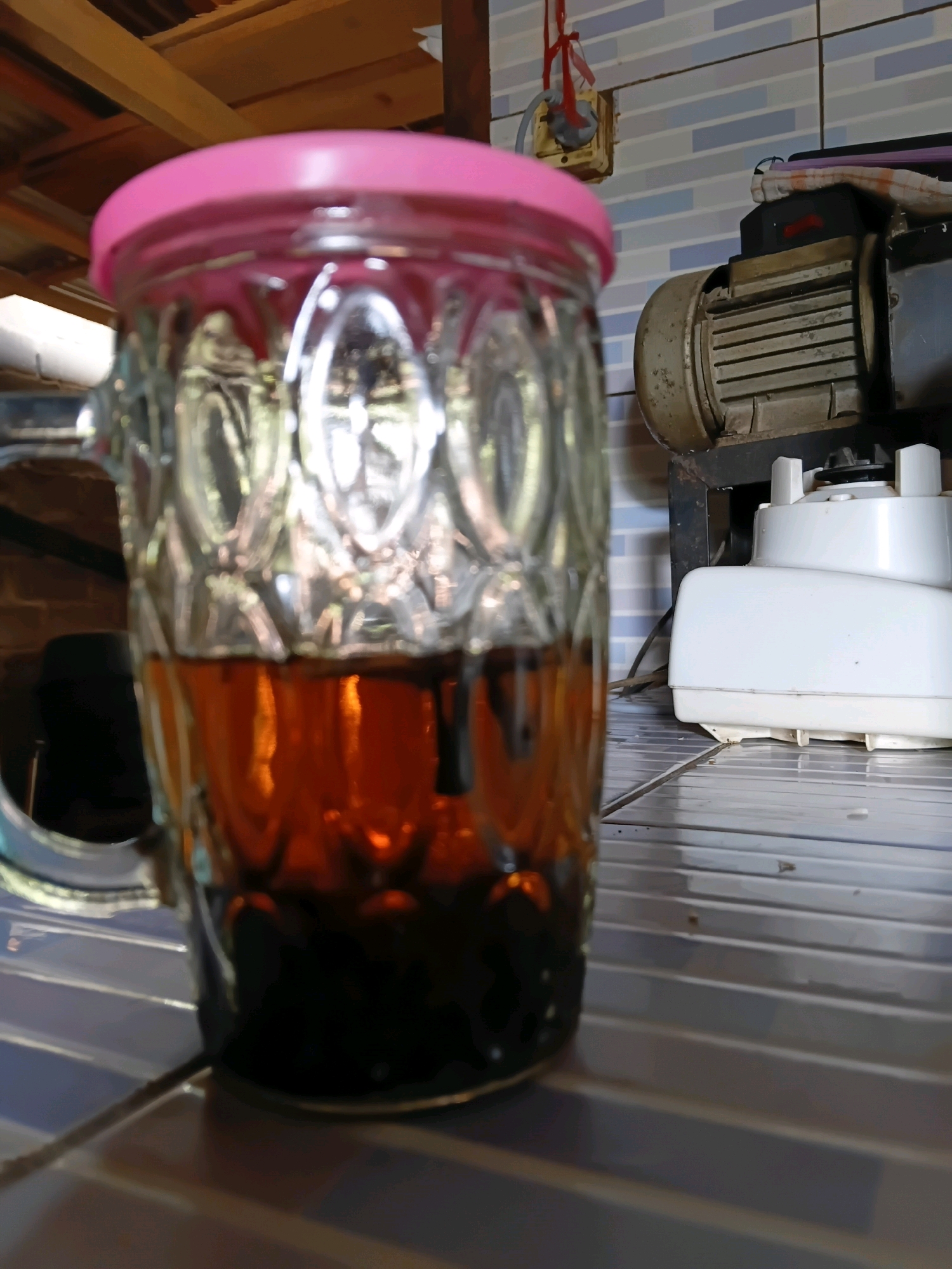 In Margo Rukun, Indonesia, a glass mug filled with a dark beverage sits on a tiled surface. The mug has a patterned glass design and a pink lid. To the right of the mug, a white blender is visible, and behind it is a large, industrial-looking motor with a metal casing. The background shows a tiled wall and wooden beams overhead. The overall scene suggests a domestic or workshop setting.