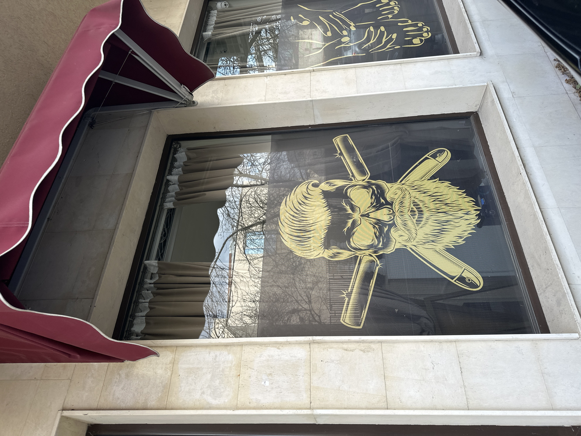 The scene depicts the exterior facade of a commercial establishment in Sofia, Bulgaria. The building features light-colored, possibly travertine, stone or tile cladding. A large display window on the left is partially covered by a burgundy-colored, scalloped awning with white trim, extended above the frame. Adhered to the window glass is a prominent gold-colored vinyl decal portraying a stylized skull with a distinct pompadour hairstyle, a full beard, and two straight razors crossed behind its head, consistent with barbershop branding. Reflections in the glass reveal bare tree branches, sections of other buildings, and the sky. Beige curtains are visible inside the premises through the transparent portions of the window. A vertical stone pillar separates this window from an adjacent, narrower window section to the right, which displays a gold decal depicting two stylized hands with palms facing upwards. A small white rectangular sticker with the number "103" is affixed to the stone pillar. The lower section of a black vehicle is faintly reflected in the bottom right corner of the image. The ground-level base of the building is also clad in the same light-colored stone.