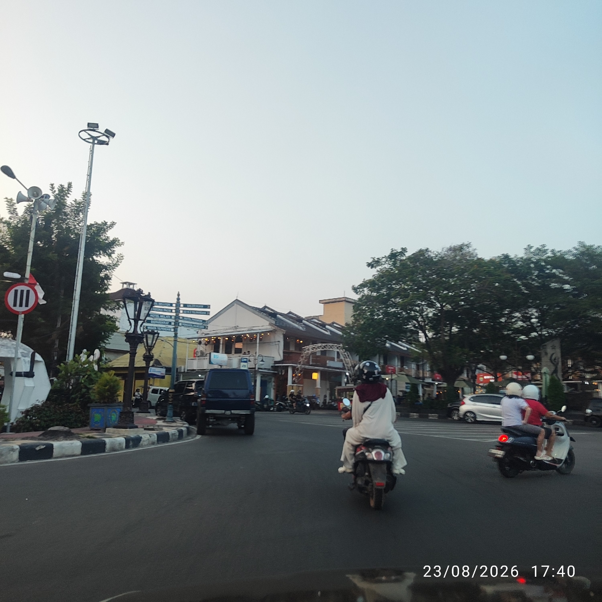 The image depicts a street scene in Tanjung Pandan, Indonesia, likely during the late afternoon given the timestamp of 17:40. The focus is on a street with vehicles and buildings.
In the foreground, a dark asphalt road curves to the left. The road is bordered by a raised sidewalk with a black and white striped pattern along its edge. To the left of the road, there is a white structure, possibly a kiosk or shelter, with greenery behind it. A tall pole with a circular light fixture and three square lights is visible on the left side of the image, along with a traffic sign indicating a prohibition with a vertical bar.
Several people on motorcycles are present. One motorcycle in the middle ground is ridden by a person wearing a white abaya and a maroon headscarf, with another passenger behind them also wearing white. Further to the right, two individuals are on a scooter; one is wearing a white shirt and the other a red shirt, both with white helmets.
In the background, a row of multi-story buildings with a traditional architectural style lines the street. These buildings feature arched windows, balconies, and decorative lights. Some of the buildings appear to be shops or restaurants. Trees are visible in the background, adding to the urban greenery. A dark blue SUV is parked on the right side of the street. The sky is clear and light blue, suggesting good weather. The timestamp at the bottom right corner reads "23/08/2026 17:40".