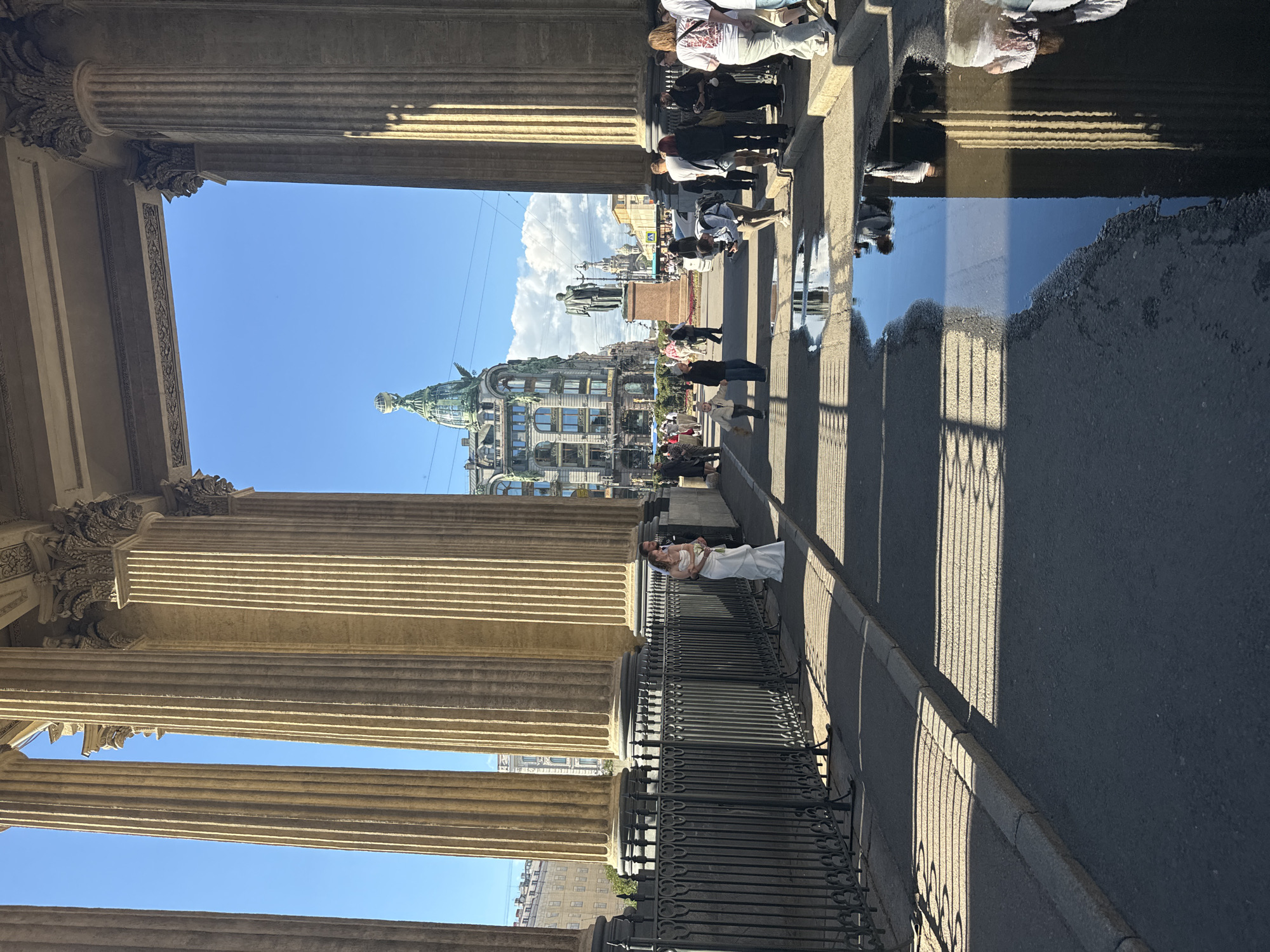 The image depicts the Kazan Cathedral in Saint Petersburg, Russia. In the foreground, large Corinthian columns cast long shadows on the pavement. A black wrought-iron fence runs along the side of the walkway. A wedding couple, the bride in a white gown and the groom in a suit, stand embracing near the fence, illuminated by the sun. Further in the background, a body of water on the pavement reflects the columns and the sky. Beyond the cathedral's colonnade, a street scene unfolds with people walking, a statue, and a distinctive green-domed building with intricate architectural details. The sky is a clear, vibrant blue with a few scattered clouds. The overall scene captures a sunny day with a mix of historical architecture and everyday life.