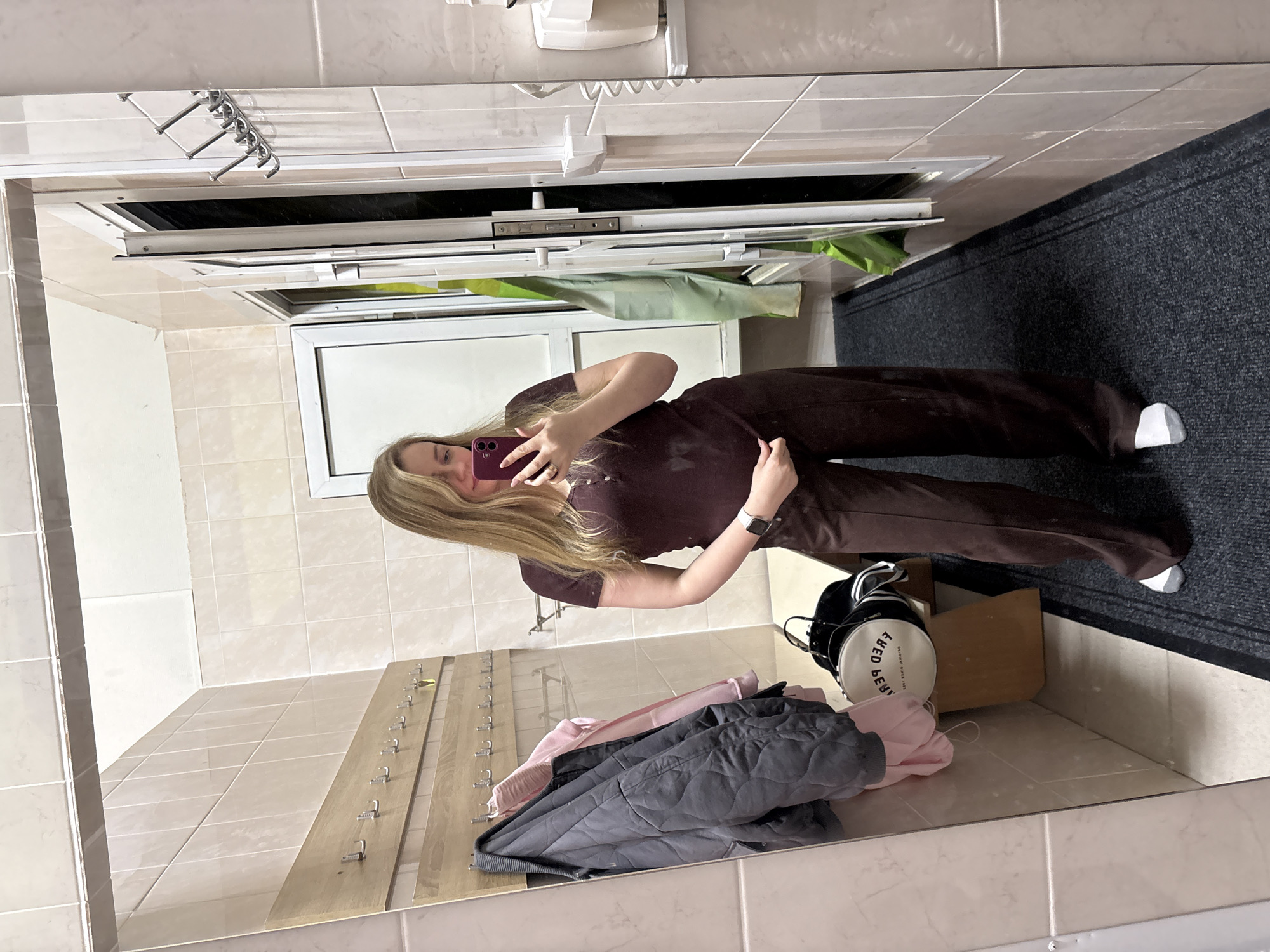 A mirror selfie is taken in what appears to be a changing room or utility space, potentially in Chaikovsky, Russia. The subject, a woman with blonde hair, is holding a red smartphone, presumably taking the photo. She is dressed in a brown matching set of a short-sleeved shirt and pants, and she is wearing a white watch. The reflection shows a tiled room with a series of hooks on a wooden panel, indicating a place to hang clothes. A pile of items, including a gray quilted jacket, a pink garment, and a white cylindrical bag, sits on the floor. Next to these items is a wooden box. A dark gray carpet or mat is partially visible on the right side of the frame. The wall is tiled, and a vent or duct is above the subject.