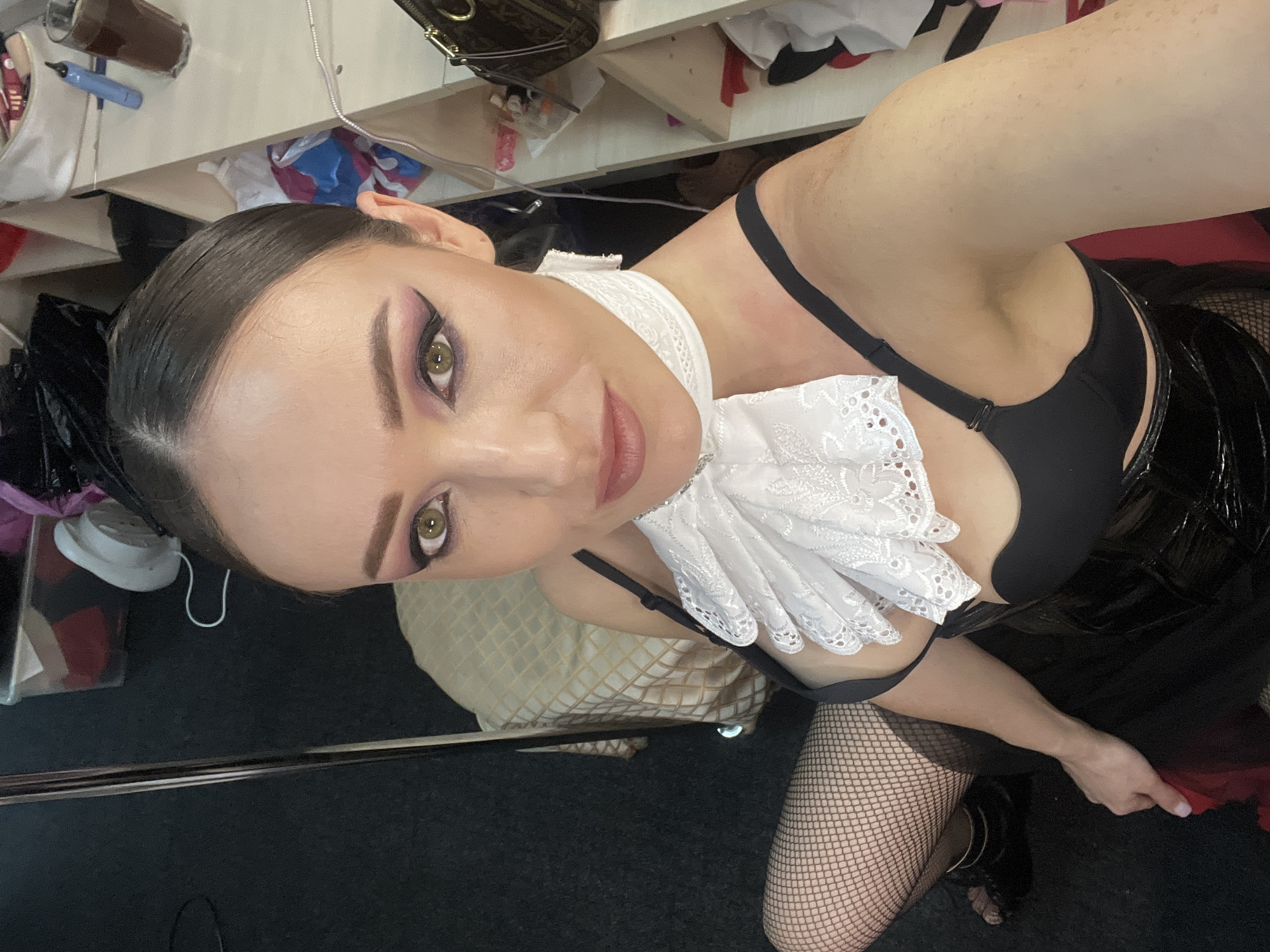 The image depicts a woman posing for a selfie indoors. She is wearing a black bra, a white ruffled collar, fishnet stockings, and black high heels. Her makeup is dramatic, featuring winged eyeliner and pink eyeshadow. The background suggests a backstage or dressing room environment, with shelves filled with various items, including clothing and personal belongings. A dark carpet covers the floor, and a chair with a patterned fabric is visible to the left. The overall impression is that of a boudoir or performance-related setting.