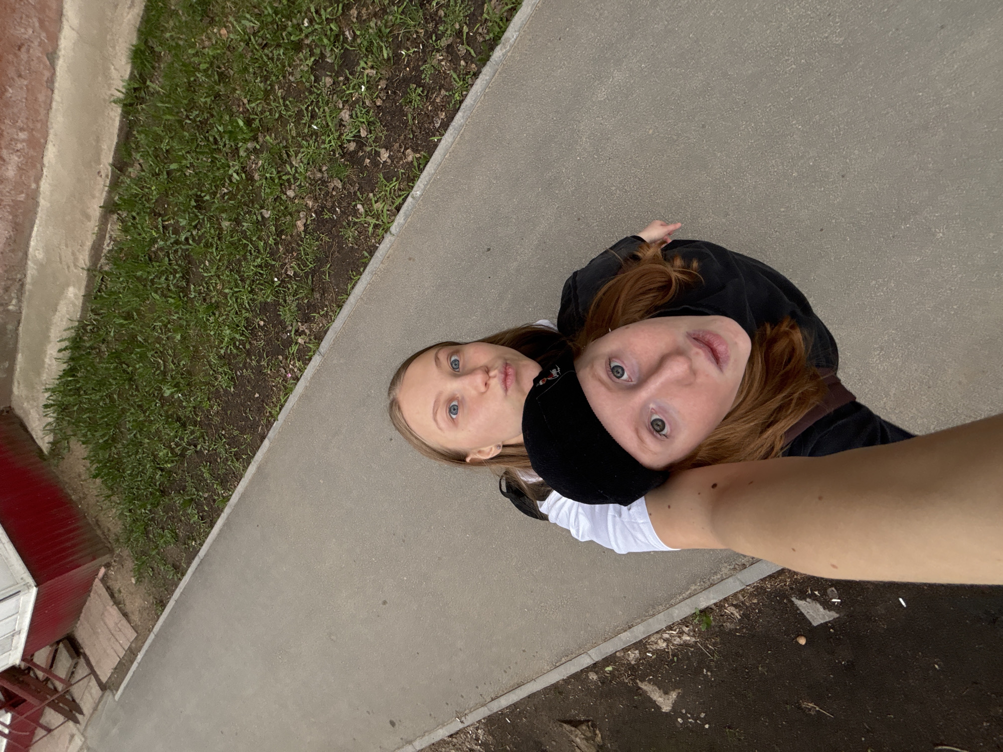 Two fair-skinned young women are posing for a selfie outside in Perm, Russia. The image, taken from a high angle, shows the women standing on a grey concrete surface, likely a sidewalk. To the left, there is a strip of grass adjacent to the sidewalk, bordered by a building with a red roof. The woman on the left has blonde hair and blue eyes. The woman on the right has red hair and is wearing a black hat. Both women are looking up at the camera. The arm of the person taking the photo is visible in the lower right corner.