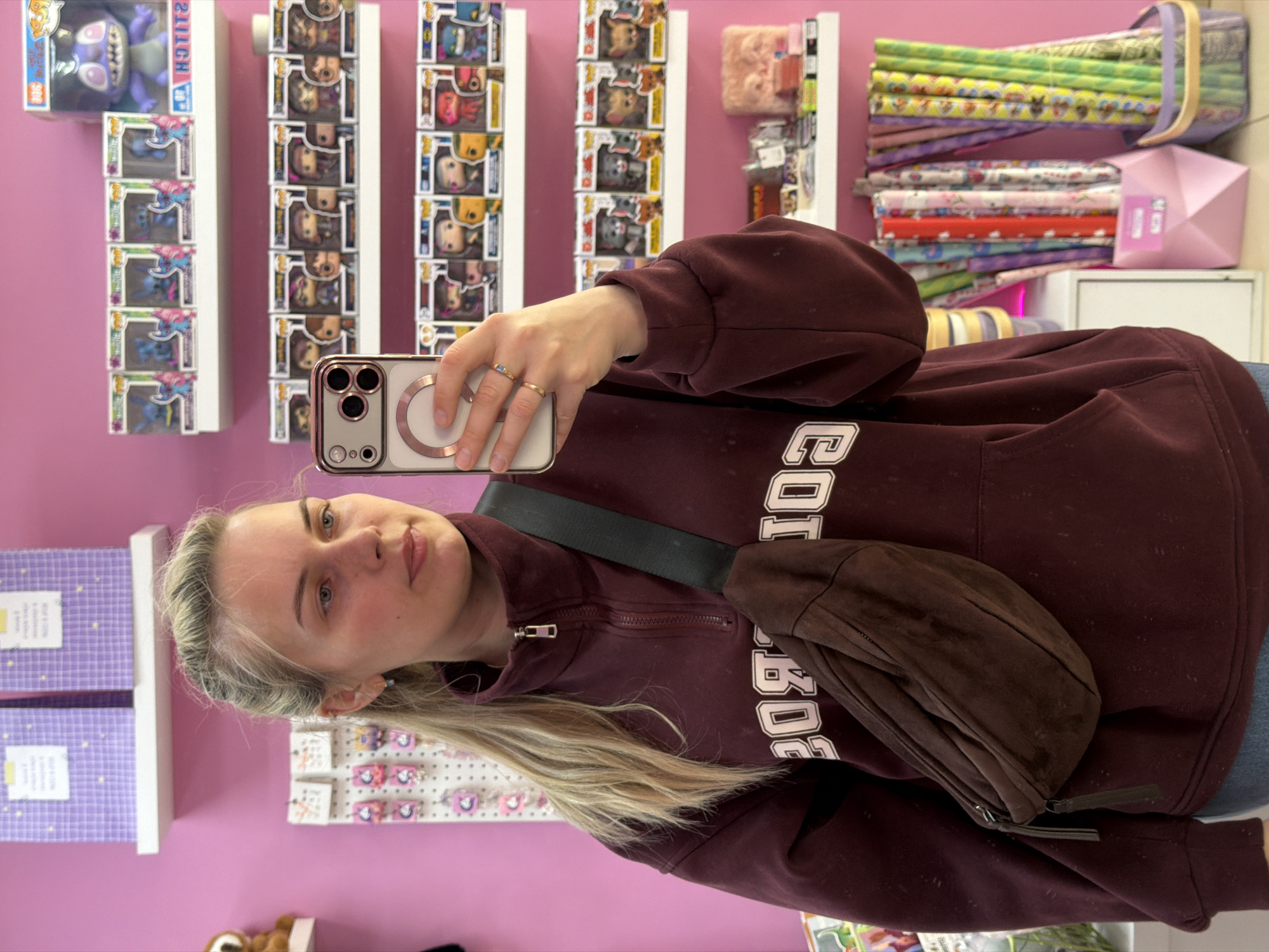 A woman with blonde hair takes a selfie in a retail environment. She is wearing a maroon sweatshirt with white lettering, and a brown suede fanny pack across her body. She is holding an iPhone to take the picture. The background shows shelves stocked with various items, including Funko Pop figures, fabrics, and other merchandise. The walls are light pink. Given the presence of distinctive retail items, it is plausible this scene takes place in a gift or souvenir shop. The location being Chaikovsky, Russia cannot be forensically confirmed based on the provided image content alone.
