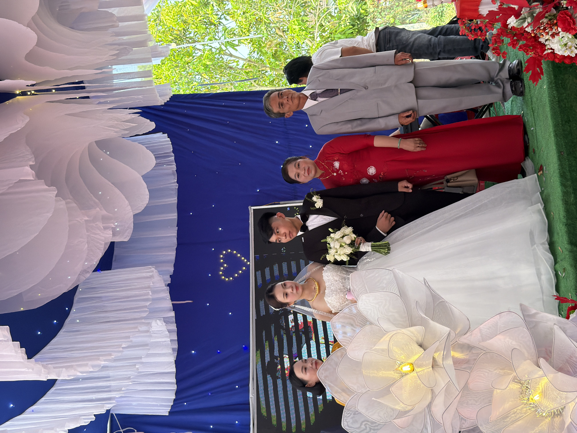 This media file captures a wedding celebration, likely held in Tay Son Commune, Vietnam. The scene is set under a tent or canopy, with natural light filtering in from above, indicating it's daytime.
In the foreground, a bride and groom are central, posing for a photograph. The bride wears a white, sleeveless wedding gown with a delicate gold necklace and veil, holding a bouquet of white flowers. The groom, dressed in a black suit, stands beside her, his hand gently resting on hers, also holding the bouquet. To their right, an additional woman, possibly a bridesmaid or guest, is partially visible, wearing a white dress adorned with large, intricate white floral embellishments that also feature small lights.
Behind the couple, a blue backdrop draped with tiny white lights mimicking stars provides a festive setting. A heart-shaped light decoration is visible on the blue backdrop to the left of the couple. Further back, an older man in a light grey suit and tie stands alongside an older woman wearing a traditional vibrant red Vietnamese dress (likely an Ao Dai) with white floral embroidery. These individuals are likely parents or close family members of the couple. Another man in a white t-shirt and dark pants is partially visible behind the man in the grey suit.
The ceiling of the event space is elaborately decorated with voluminous white fabric drapes, creating a cloud-like effect, some interspersed with small, warm lights. On the right side of the frame, red floral arrangements and greenery add splashes of color. The ground is covered with a green surface, possibly artificial grass or a carpet, creating a garden-like feel indoors. A large screen or LED display is visible behind the bridal party, showing pixelated patterns. There is no visible text in the image. The atmosphere is celebratory and formal, characteristic of a wedding ceremony or reception.