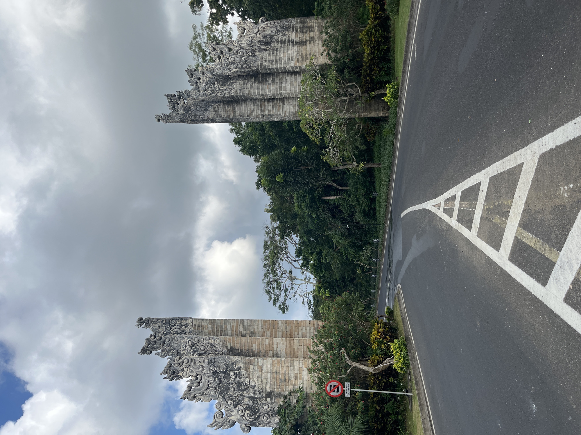 The image shows a road leading through a large, ornate stone gate in Kuta Selatan, Indonesia. The gate is made of intricately carved stone and is flanked by lush greenery. The road is paved and has white lane markings. The sky is cloudy. There are no people visible in the image. The gate appears to be a significant landmark, possibly an entrance to a temple or a residential complex. The overall impression is one of grandeur and tropical beauty.