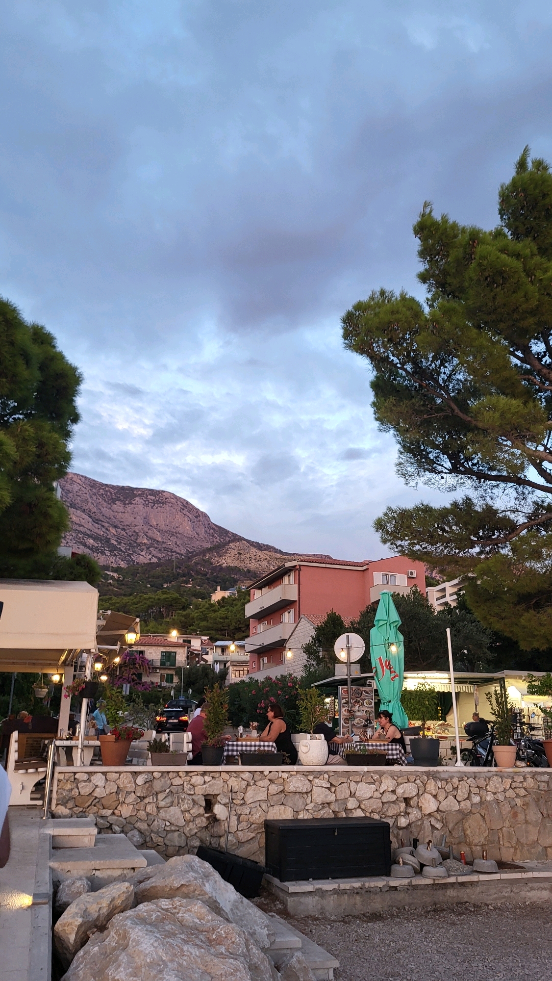 The image depicts an outdoor restaurant scene in Općina Tučepi, Croatia, at dusk. In the foreground, large, rough stones are piled on a paved surface. A grey, rectangular storage chest sits on a concrete ledge behind the stones. Further back, a stone wall borders a raised patio area where several tables are set up with checkered tablecloths. Three women are seated at these tables. One woman in the center, wearing a black dress, is looking to her right. Another woman to her left, also in a black dress, is looking down. A third woman, partially visible, is seated at a table further to the right.
Behind the patio, a pink building with balconies stands prominently, with several other buildings visible in the background, suggesting a coastal town nestled against a mountain. The mountain itself, with its rugged, rocky surface, dominates the background landscape, appearing purple-tinged under the cloudy sky. Lush green trees, likely pine trees, frame the scene on the left and right sides of the image, their branches reaching towards the sky. The sky is filled with a mix of grey and white clouds, indicating overcast conditions or the transition from day to night. A bright green umbrella is visible over one of the tables. The overall atmosphere suggests a relaxed evening setting.