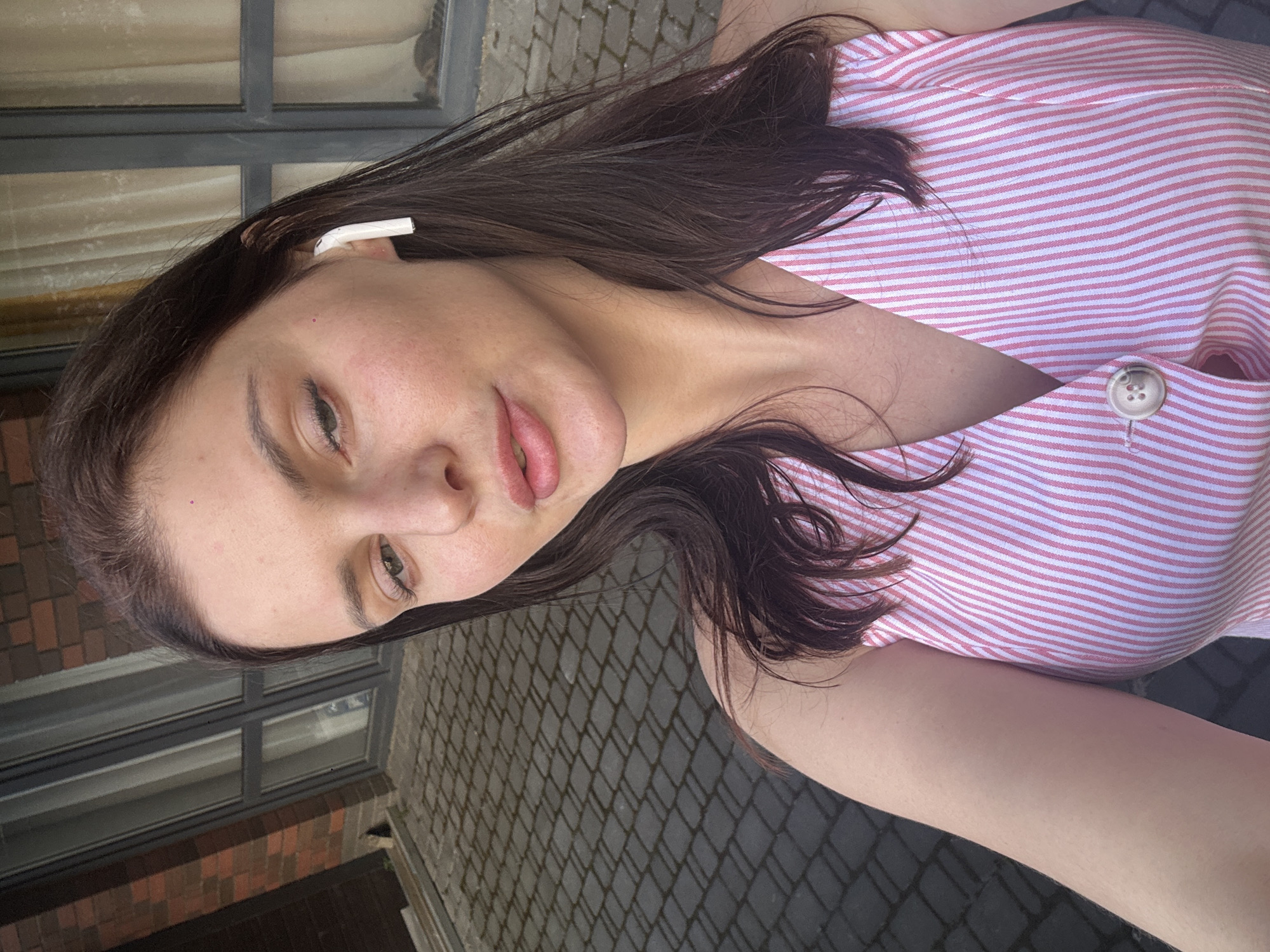 A young woman is captured in a close-up selfie, her face angled slightly towards the camera. She has long, dark brown hair that falls around her shoulders. A white wireless earbud is visible in her right ear. She is wearing a pink and white striped sleeveless top with a V-neckline and a single button closure. The background suggests an outdoor setting with a textured grey paved ground and a brick wall with windows to her left and right. The lighting indicates it is daytime.