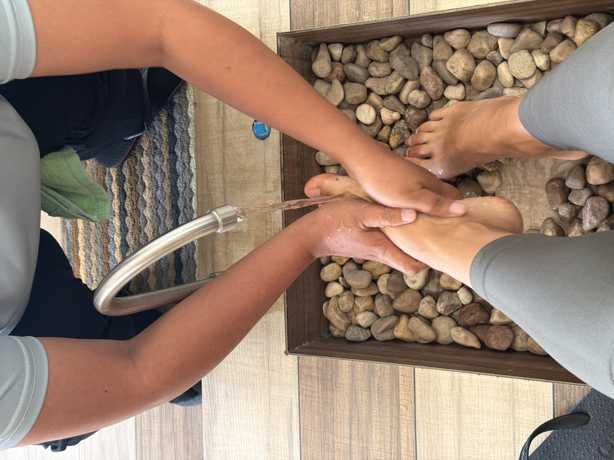 A person with brown skin is washing or massaging the feet of another person with brown skin. The feet are placed in a wooden tray filled with smooth, round river rocks. Water is being poured from a silver faucet onto the feet. The person whose feet are being washed is wearing gray leggings and sitting on a gray and brown striped rug. The setting is indoors, on a wood-grain floor. It is likely that the scene is a spa treatment located in General Luna, Philippines.