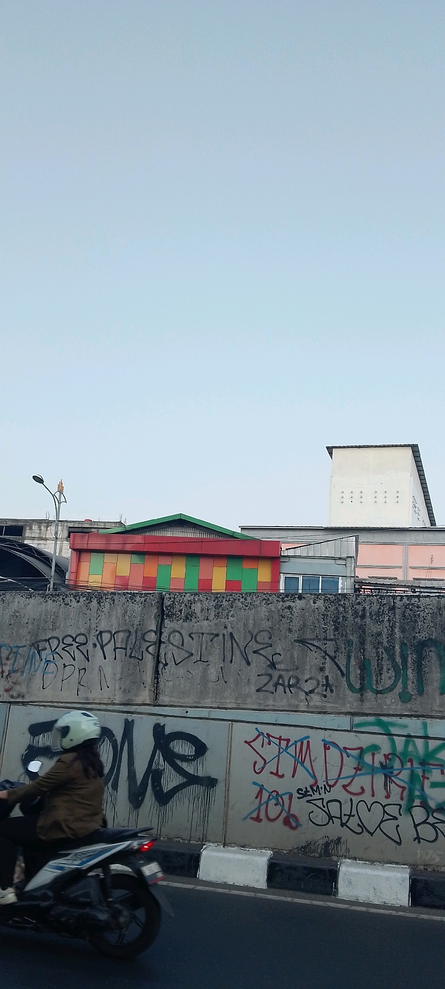 An exterior, medium shot captures a street scene in Bekasi, Indonesia, under an overcast sky. The frame is divided horizontally by a concrete barrier covered in graffiti. In the foreground, a person on a white and blue motorcycle with black accents is riding away from the camera. The rider wears a brown jacket and a white helmet. The motorcycle's rear light is on. The concrete barrier is adorned with various graffiti tags in black, red, and green spray paint. Prominent text includes "FREE PALESTINE," "DPR," "ZARQI," "ONE," and "SIMDZIRE." Above the barrier, a colorful building with red, yellow, and green panels is visible, suggesting it might be a commercial establishment or entertainment venue. Behind this building, other structures with flat roofs are present, including a white building with a sloped roof and several small, regularly spaced circular openings. A lamppost with a bent neck stands on the left side of the frame. The road surface in the foreground is dark asphalt, bordered by a concrete curb painted with alternating black and white stripes.