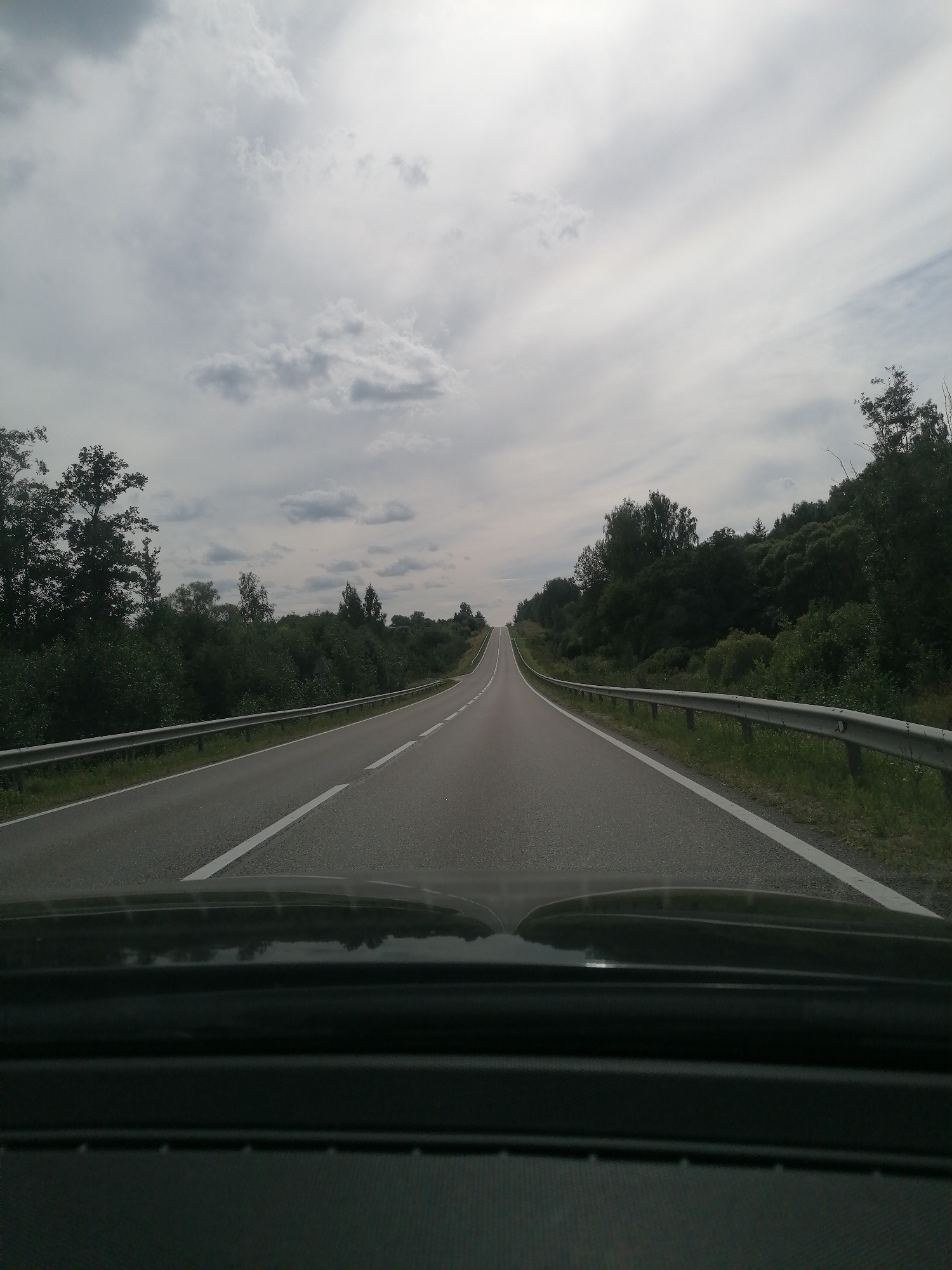 The image shows a straight, two-lane asphalt road with white lane markings, likely a highway or main road, curving slightly to the right. The road is bordered by a metal guardrail on both sides. Lush green trees and bushes line the sides of the road, forming a dense forest on the right and a more sparse collection of trees on the left. The sky is overcast with light gray clouds, indicating a possibly cloudy or overcast day. The perspective is from inside a vehicle, with the dashboard visible in the foreground. The scene appears to be in a rural or semi-rural area, possibly on the outskirts of Randene, Latvia, given the natural landscape and road conditions. No people or specific actions are depicted in the image.
