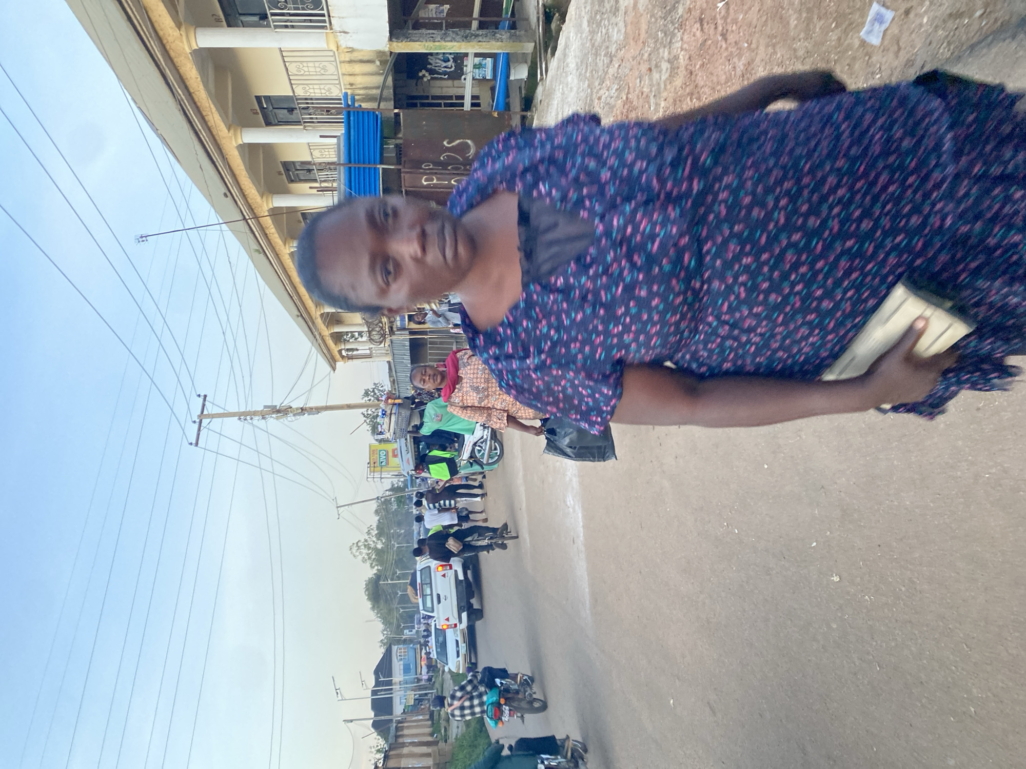This image captures a street scene in Gura Top, Nigeria, likely during the late afternoon or early evening. The foreground is dominated by a woman walking towards the camera, her face turned slightly away. She is wearing a dark blue dress with a repeating pattern of small pink and green dots. In her right hand, she holds a light-colored clutch or wallet. Her expression appears neutral.
Behind her, the street is bustling with activity. Several people are walking and riding motorcycles and bicycles. A white pickup truck is visible in the middle ground, as are other vehicles and motorcycles. The buildings lining the street are two-story structures with balconies and columns, suggesting a commercial or residential area. Power lines crisscross the sky, a common feature in Nigerian urban environments. The road surface is asphalt, with some debris scattered along the side. The overall atmosphere suggests a busy, everyday scene in a Nigerian city.